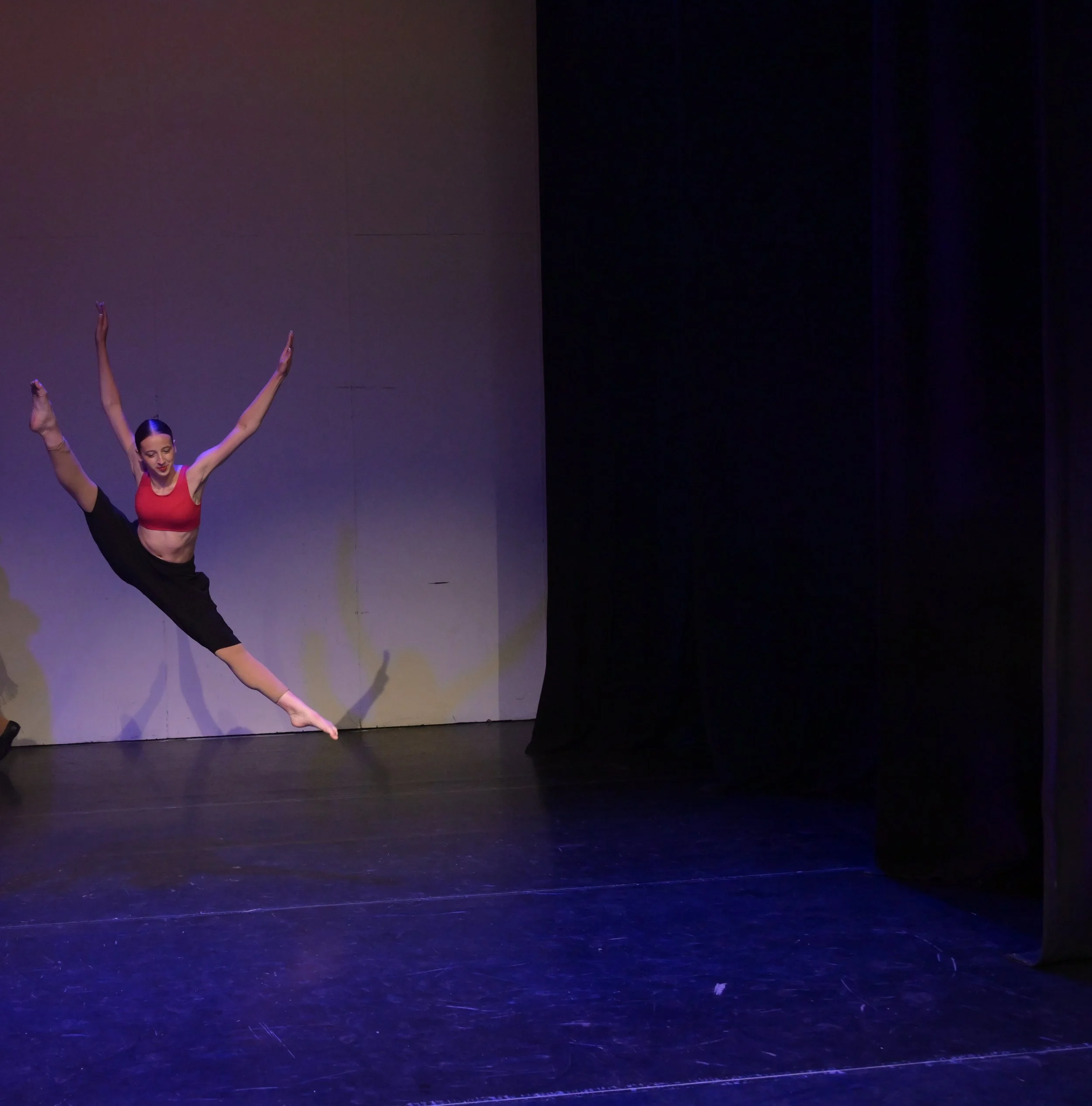 A female dancer in a red top and black skirt performing a leap on stage with a plain background and dark curtains.