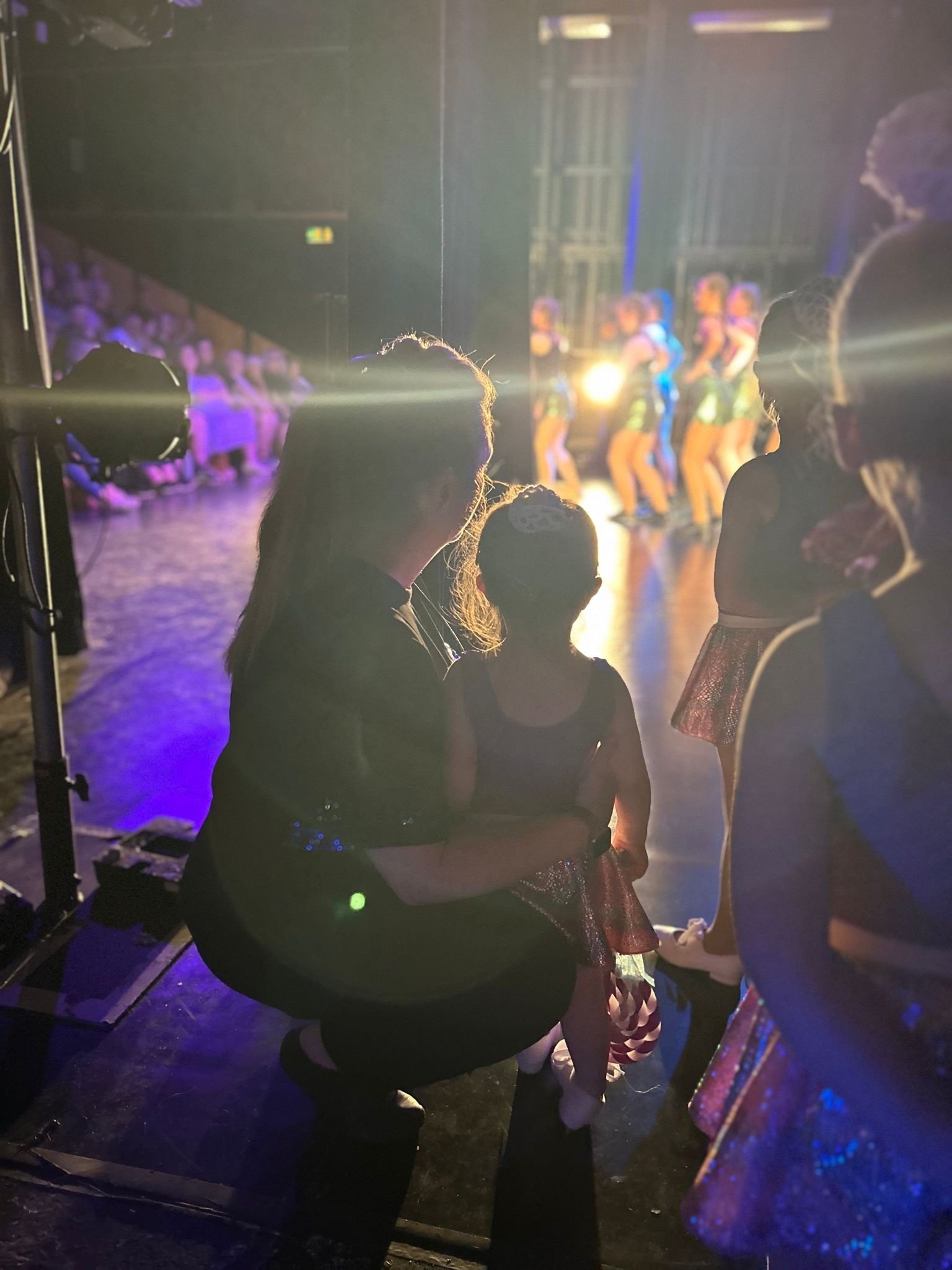 A young girl and a woman sitting backstage, watching a dance performance on stage with bright lights. Other people are visible, some standing and some sitting, with dancers performing in the background.