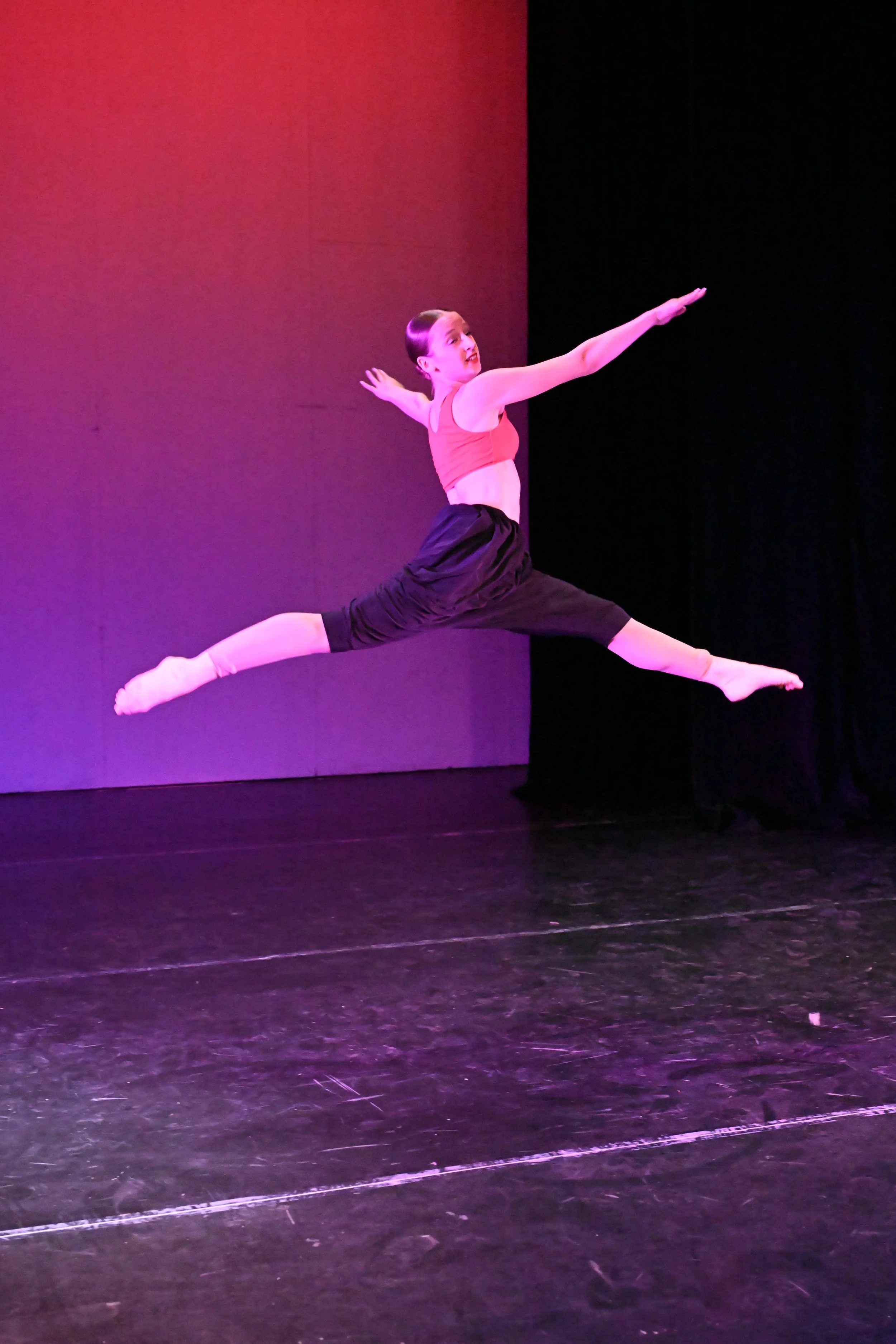 A female dancer in a pink sports bra and black pants performing a grand jeté leap on stage with a colorful lighting background.