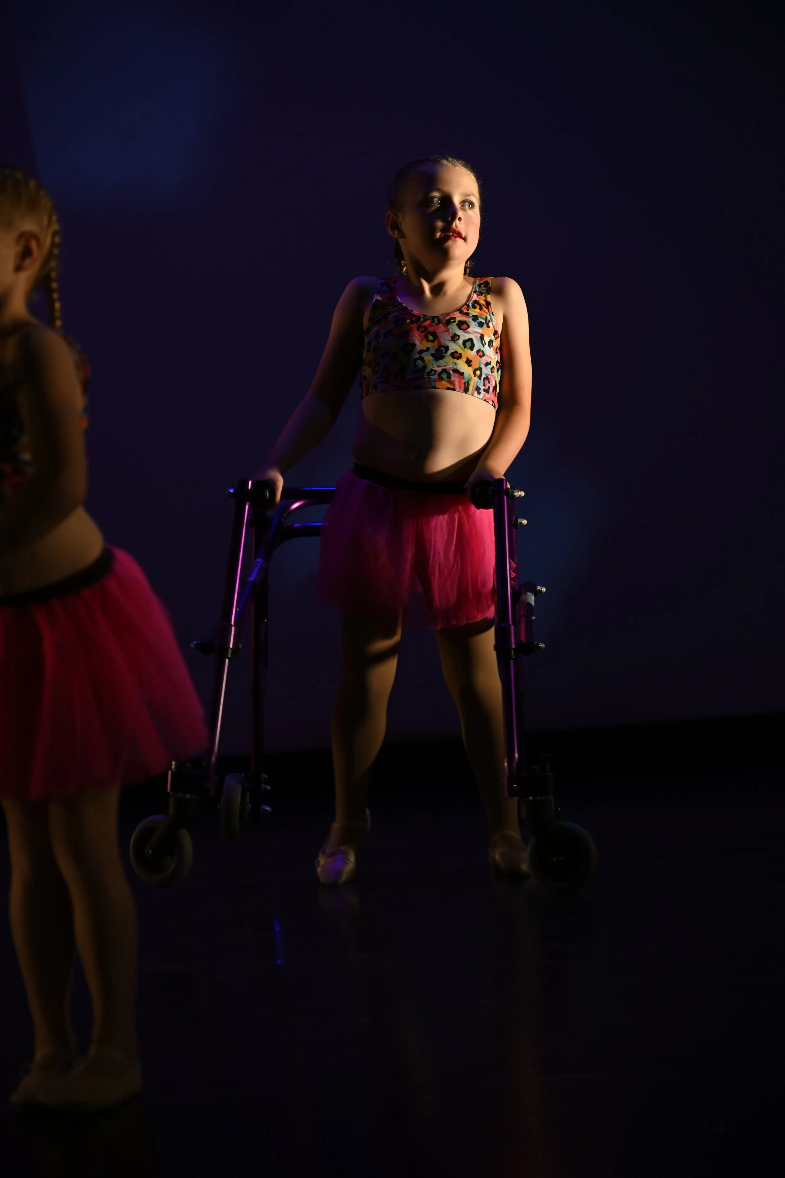 A young girl in a dance costume standing with the help of a walker on stage, lit by stage lights