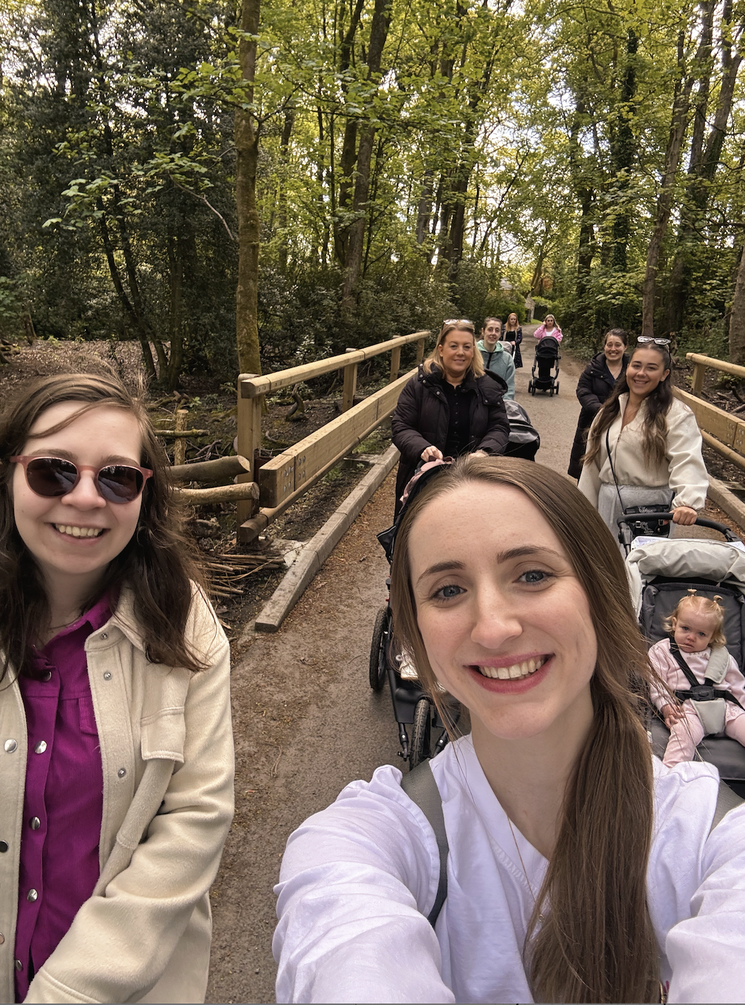 Group of women and children walking on a nature trail in a wooded area, smiling for a selfie, some individuals using strollers, surrounded by trees and greenery.