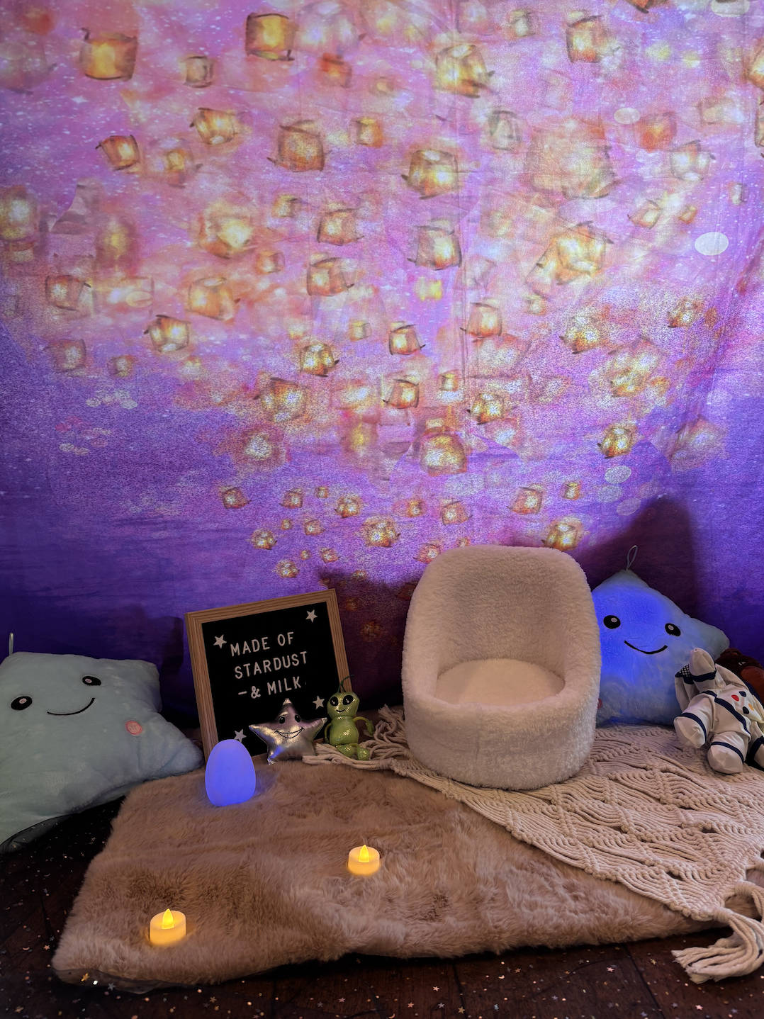 A cozy space setup with plush pillows, soft blankets, and small decorative lights. A backdrop features a pink, purple, and gold star and lantern pattern projected on the wall. There are plush toys, a sign that reads 'Made of Stardust & Milk,' and sma