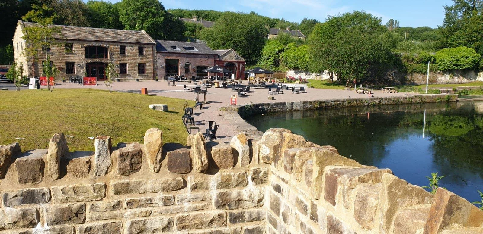 Outdoor scene with a historic stone building near a pond, surrounded by trees and park benches. Clear sky, sunny day.