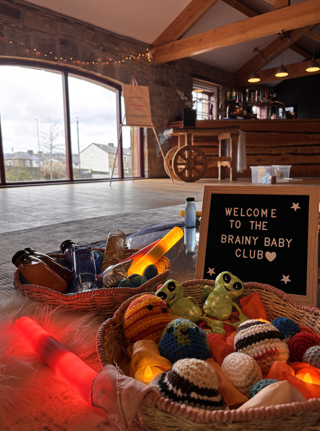 Indoor space decorated for a baby club event with baskets of knitted baby hats and plush toys, a chalkboard sign that reads "Welcome to the Brainy Baby Club," and colorful lights, with a bar area in the background and large glass doors showing an out
