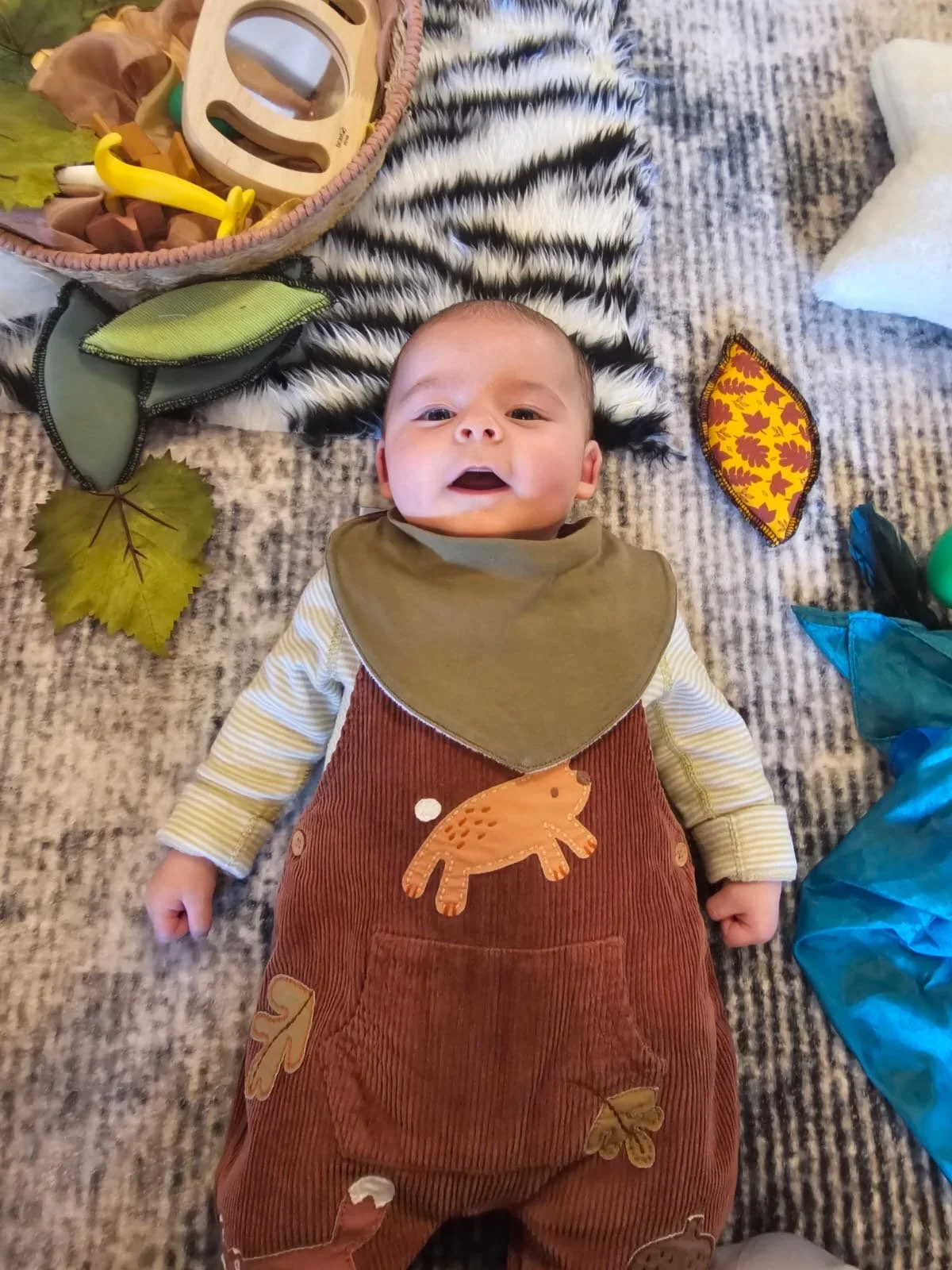 Baby lying on a striped black and white blanket on a gray carpet, surrounded by toys and artificial leaves, with a smile and wearing an orange dinosaur-themed outfit and a green bib.