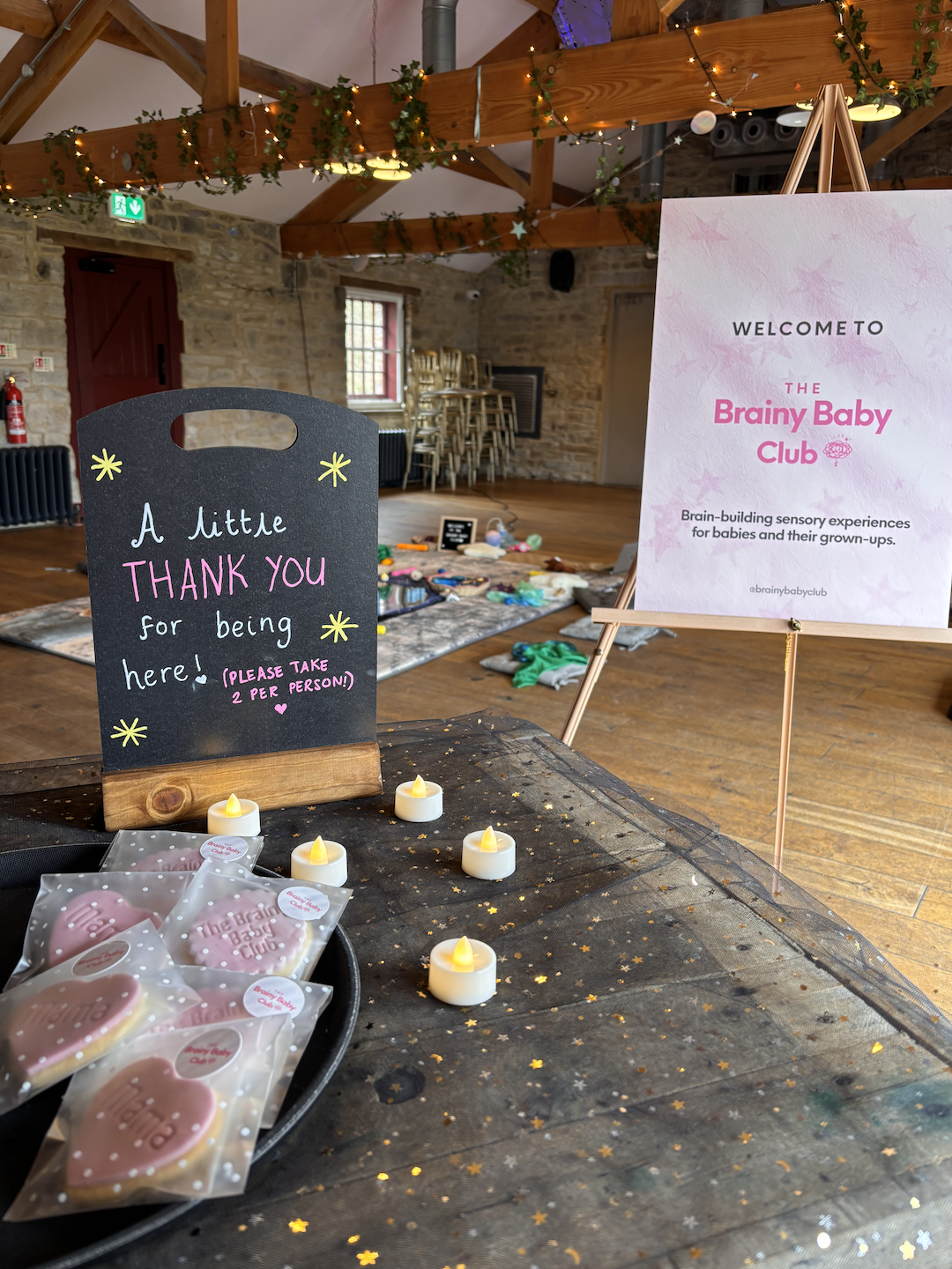 Decorated event space with a welcome sign for 'The Brainy Baby Club', small candles, pink cookies, and a board thanking attendees for being there. The room has wooden beams, a stone wall, and a play area with toys on the floor.