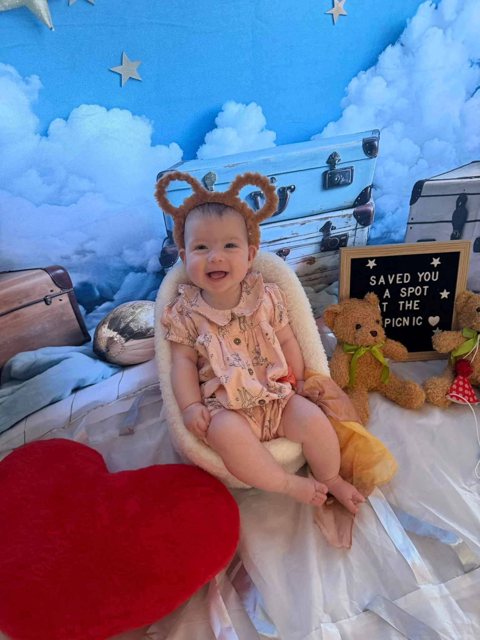 A smiling baby girl wearing a pink dress with a bunny print, sitting in a cozy chair with a fluffy white cover, surrounded by soft toys including a bear with a green bowtie, and a red heart-shaped pillow in the foreground. The background features blu