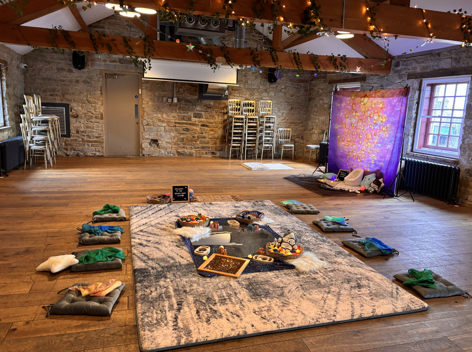 A decorated indoor space with a wooden ceiling, string lights, and a mix of chairs stacked and scattered. The walls are made of stone, with windows on the right. The floor has a large rug with various small cushions and children's toys laid on and ar