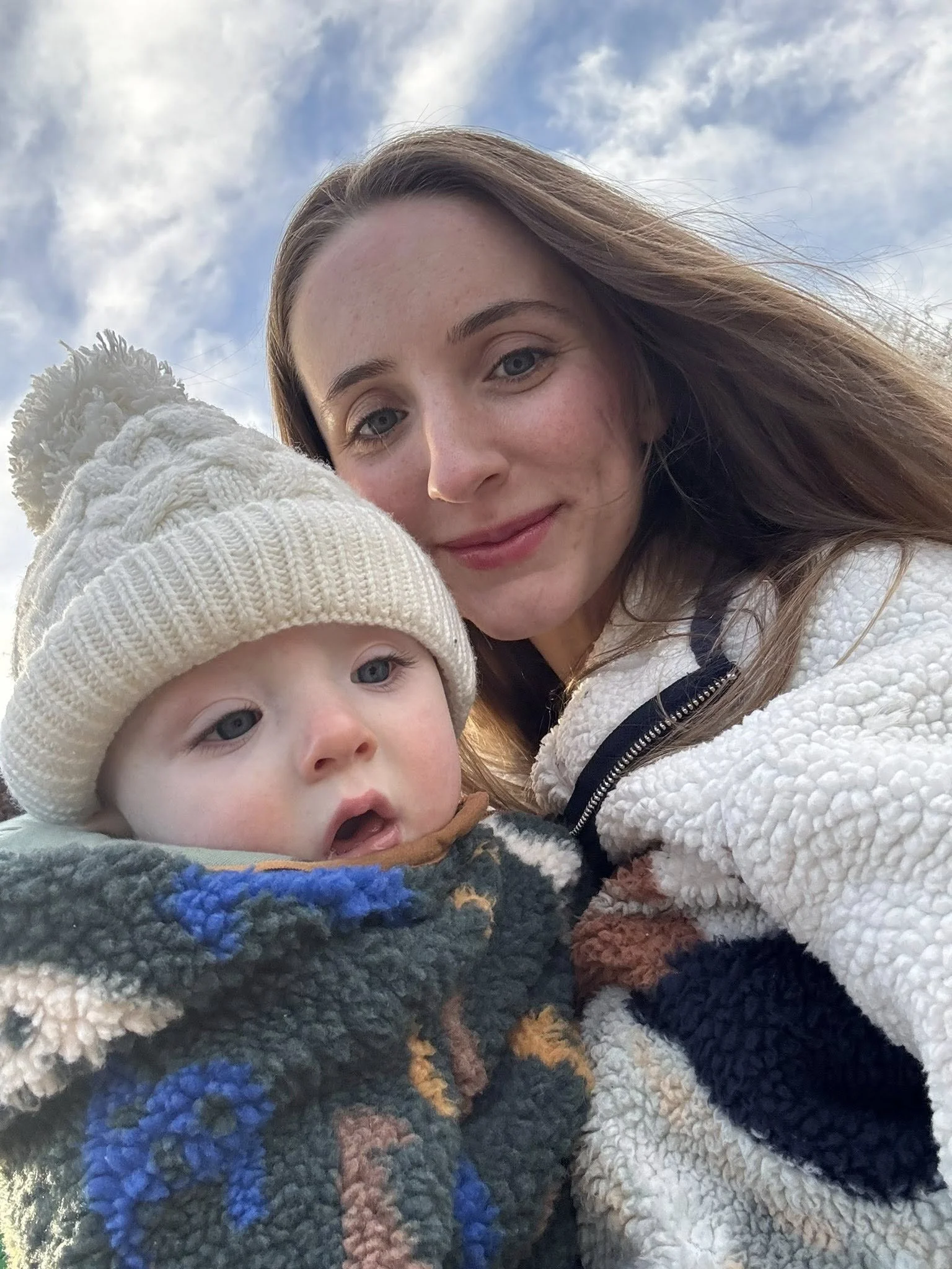 A woman and a young child taking a selfie outdoors, both wearing warm clothing. The woman has long brown hair and is smiling softly. The child, wearing a knitted hat with a pom-pom, is wrapped in a colorful fleece blanket and has a curious expression. The sky is partly cloudy.