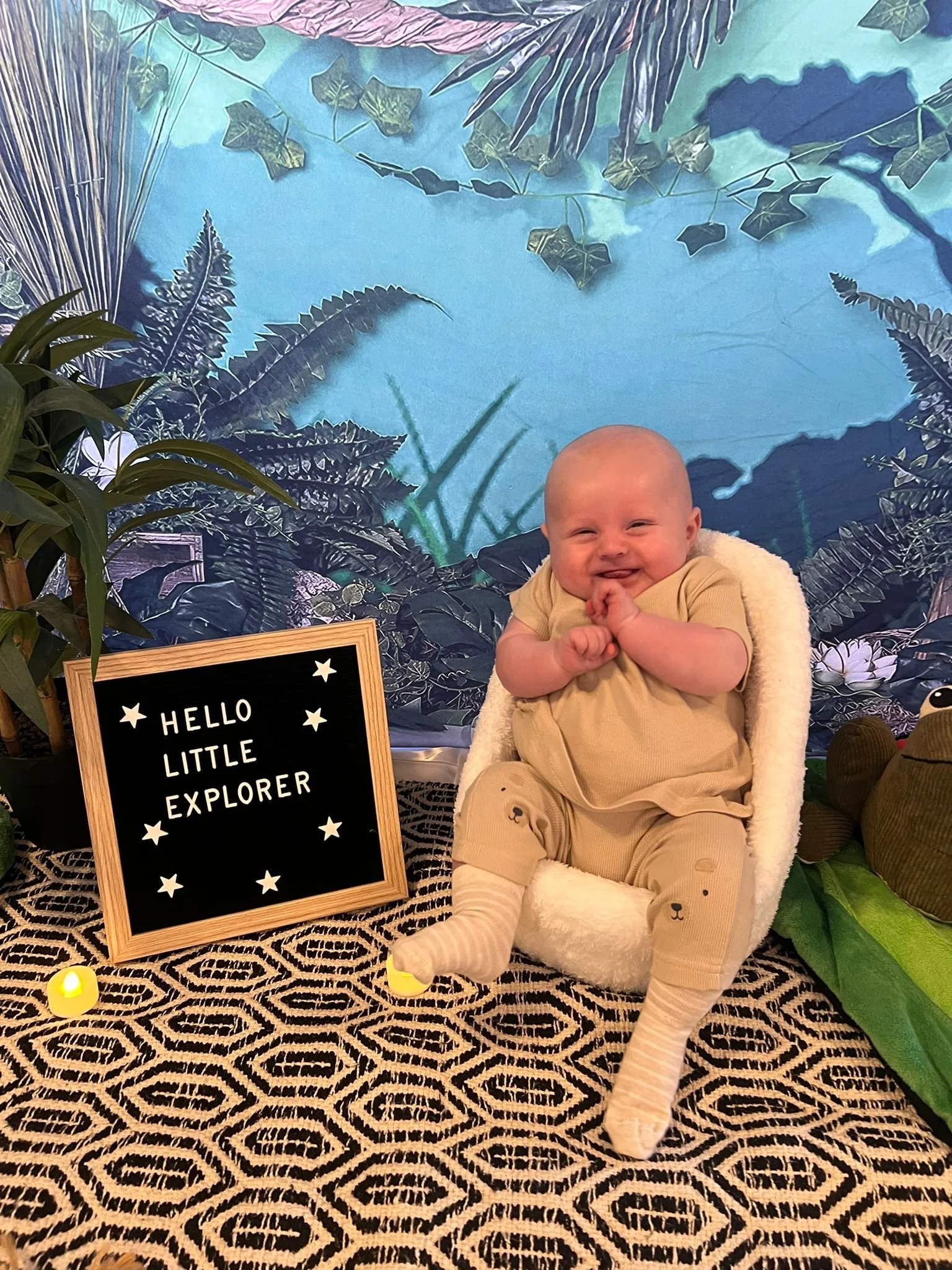 A smiling baby sitting in a plush chair with a fluffy blanket, surrounded by greenery and decorative plants, with a sign reading 'HELLO LITTLE EXPLORER' nearby.
