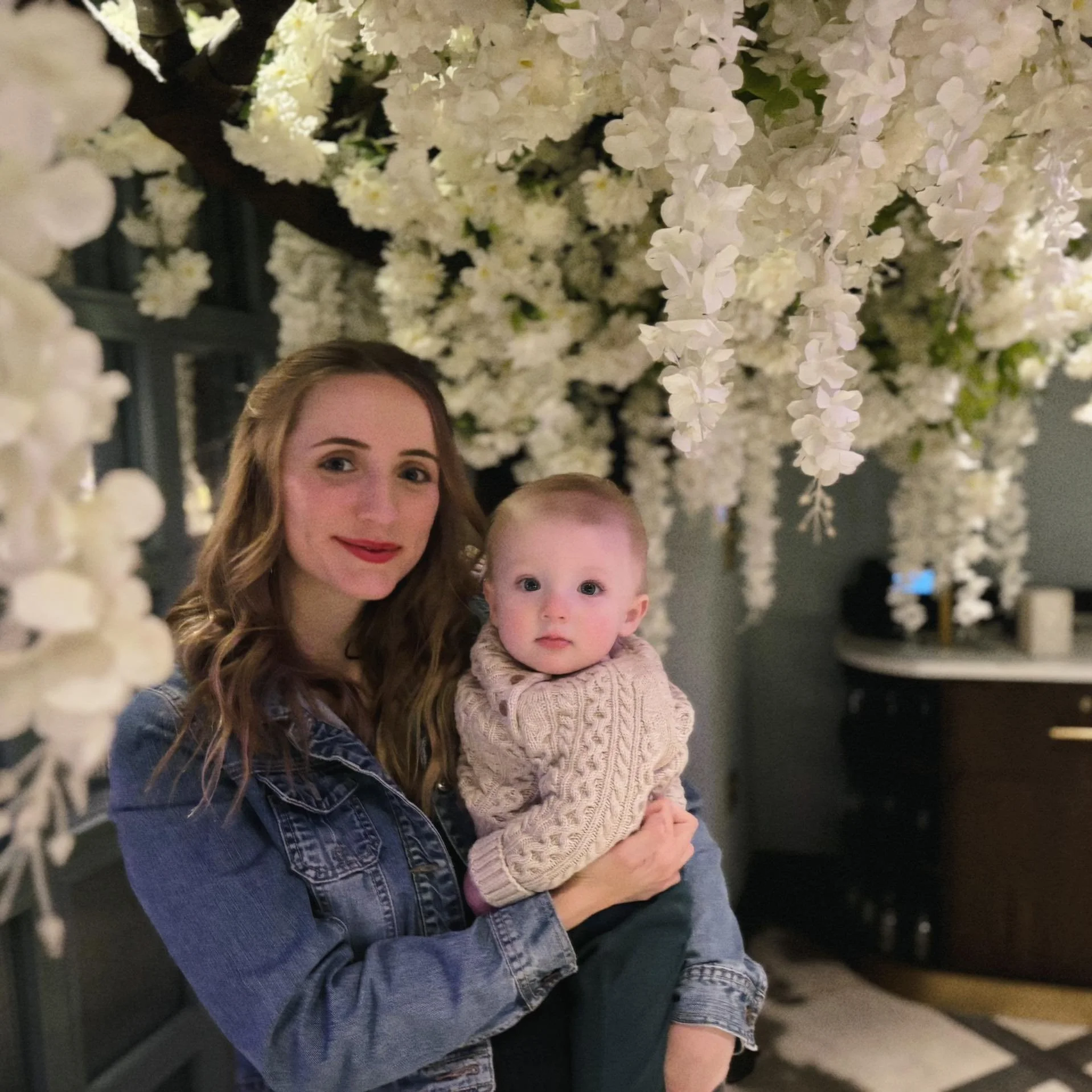 A woman with long, wavy, light brown hair holding a young child with short blonde hair, standing in front of a large, hanging display of white flowers inside a dimly lit room.