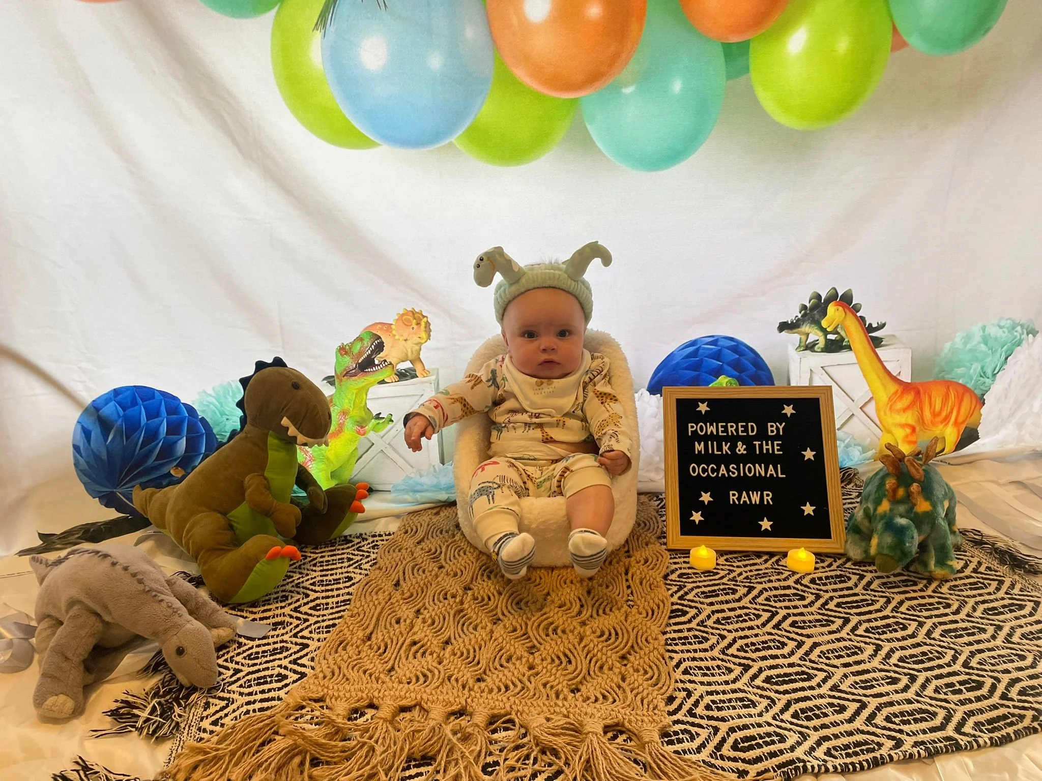 Baby sitting among colorful dinosaur and animal plush toys, with balloons overhead, on a patterned rug, and a sign that reads "Powered by Milk & The Occasional Rawr," in a playroom or party setting.