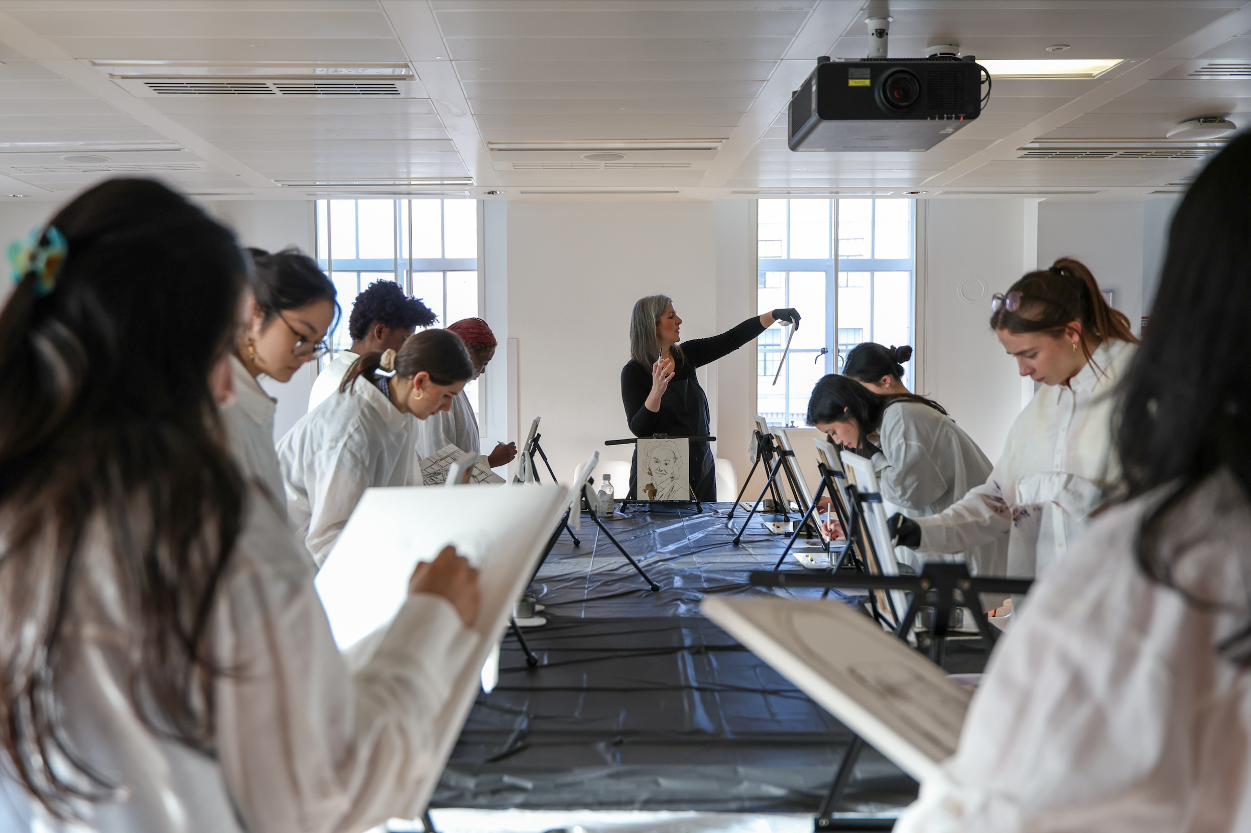 A team participating in a guided painting workshop, sitting at easels, with a female instructor standing at the front, demonstrating techniques, in a well-lit room with large windows.