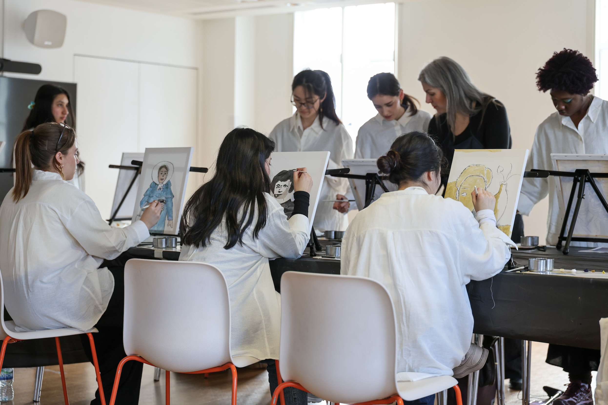 A group of people participating in a guided painting workshop, sitting and standing around tables with easels, creating portraits and artwork on canvases in a well-lit room.