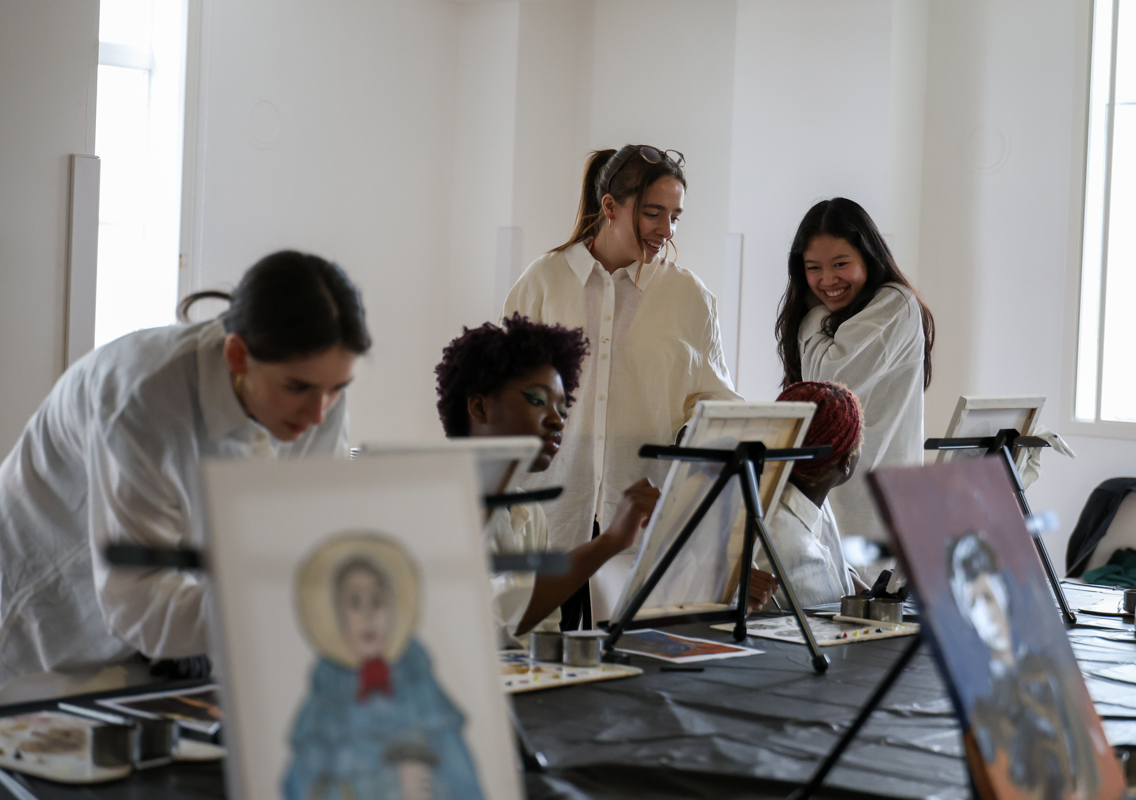 A group of women painting portraits on canvases, with some women working on their paintings and others observing and smiling at each other in a bright, indoor studio.