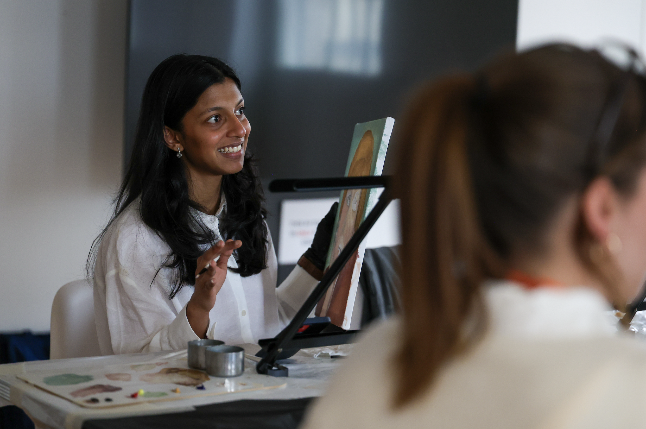 A team member sitting painting at an easel with an art palette and paintbrushes, with another person in the foreground.