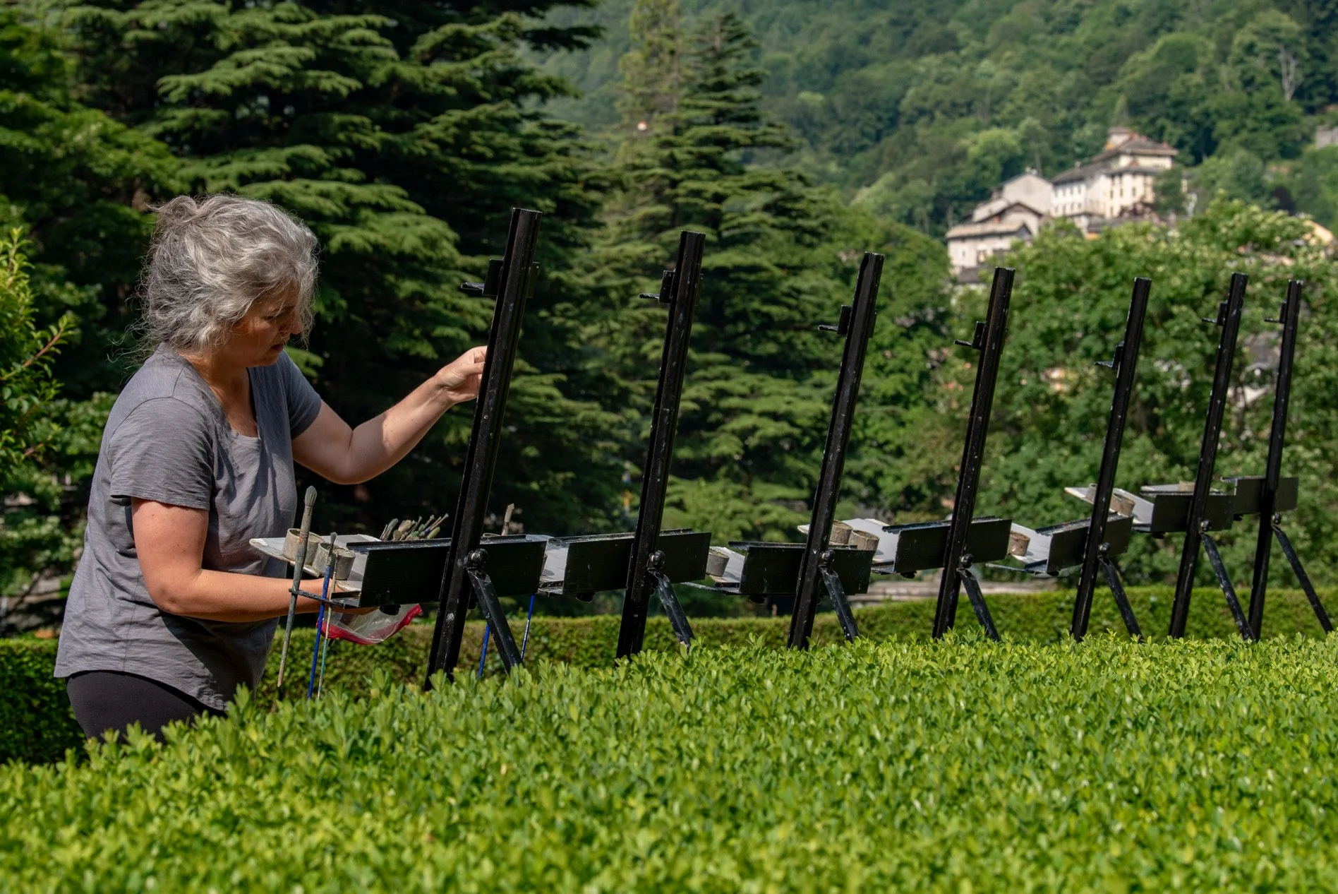 A woman painting pleinair in a green garden, standing alongside multiple easels with her palette in hand, surrounded by trees and a distant view of buildings on a hillside.