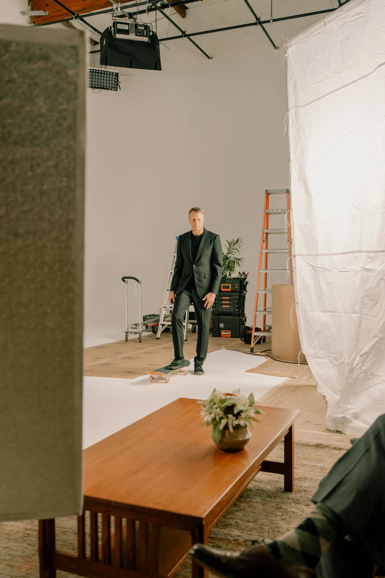 A man in a black suit standing on a skateboard in a photography studio with lighting equipment, ladders, and a white backdrop. A table with a flower arrangement is visible in the foreground.