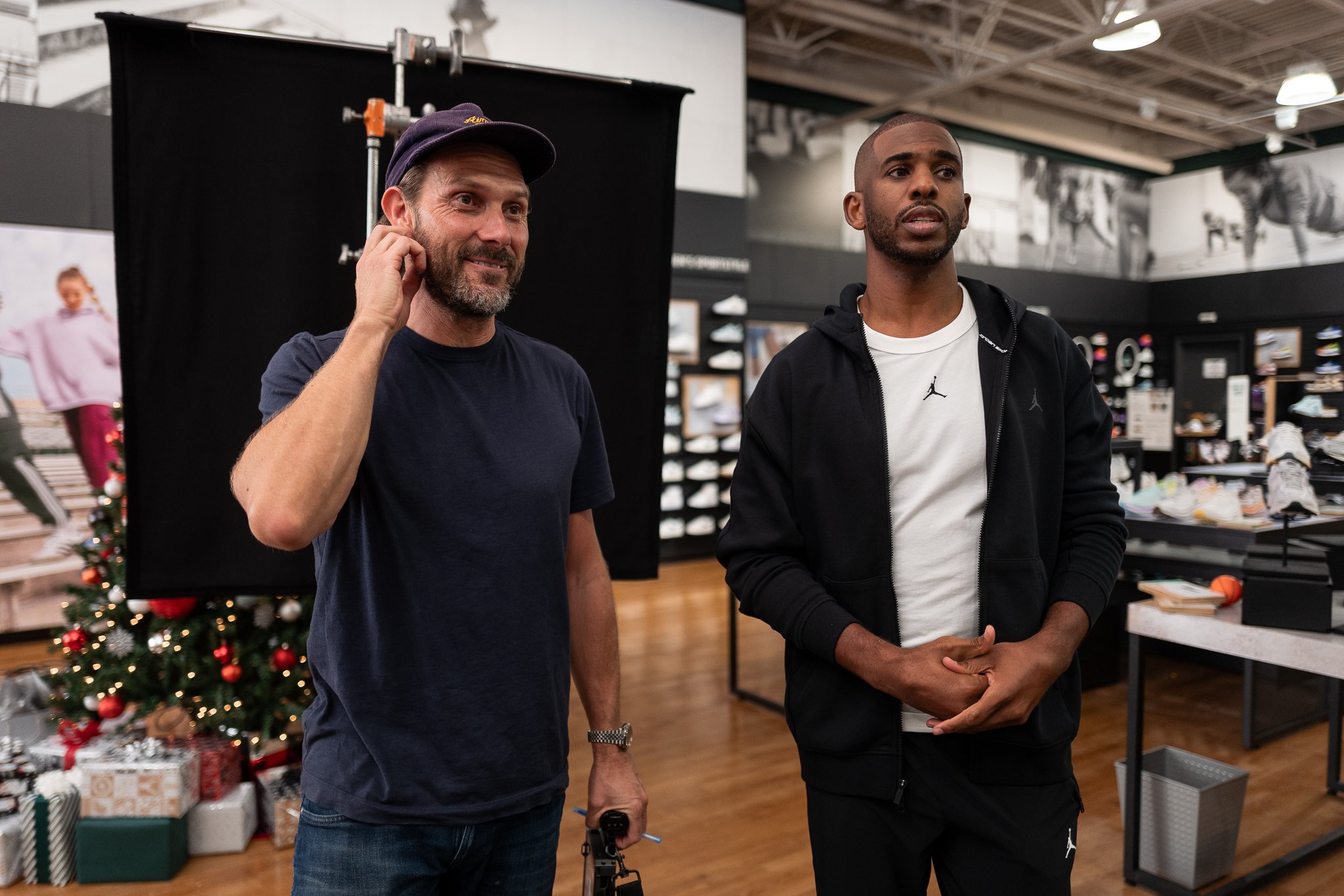 Two men standing inside a retail store, one holding a camera, with shelves of shoes and a decorated Christmas tree in the background.
