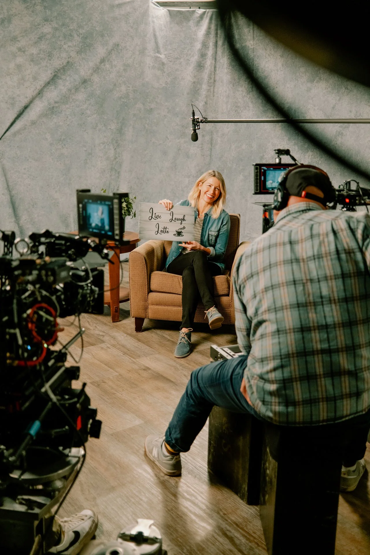 A woman sitting in a chair holding a sign that says 'Live Laugh Latte' next to a camera crew filming her in a studio setting.