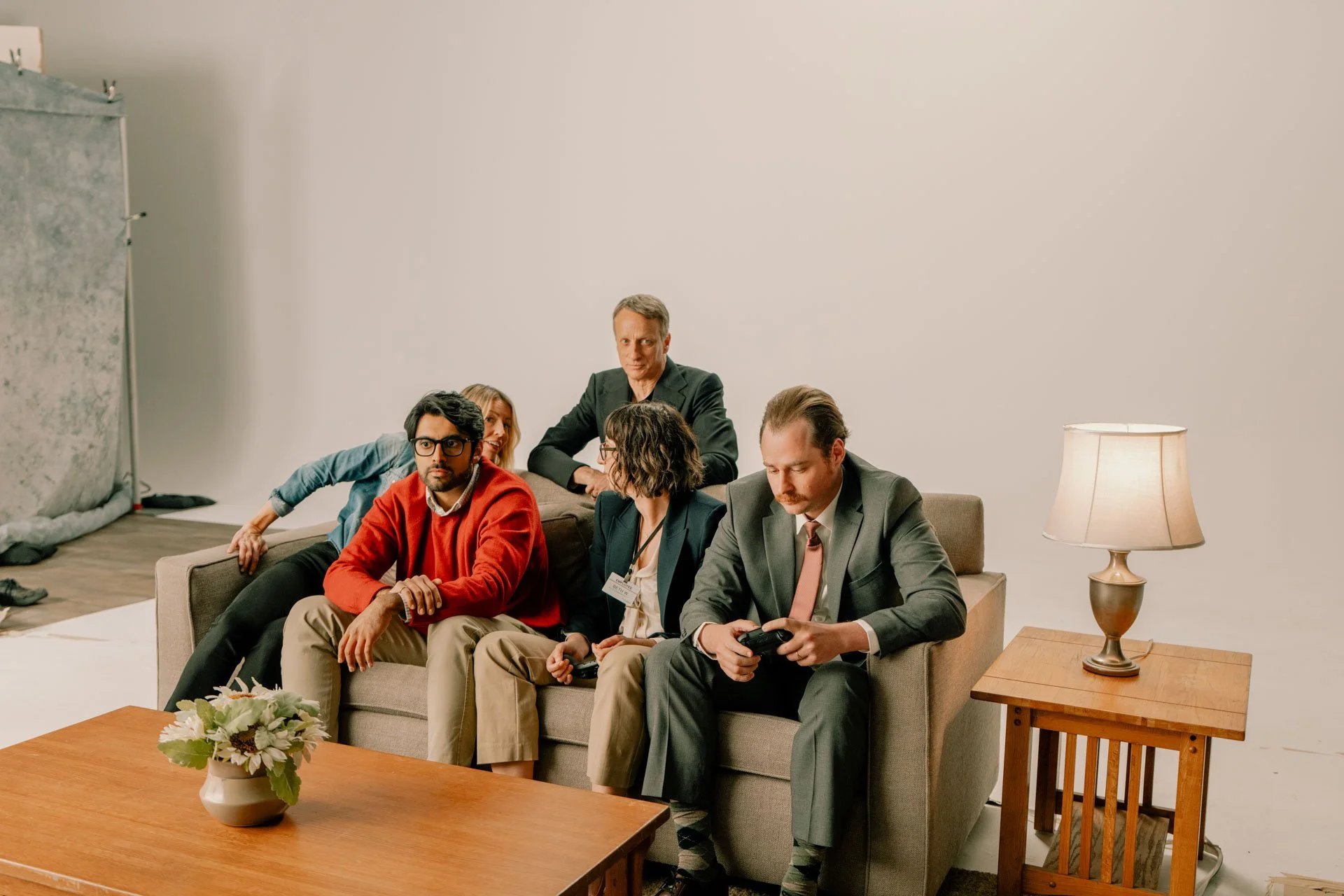 Five people sitting on a sofa in a photography studio, with a plain white background. One person is sitting on the floor, leaning forward sideways, partially behind the armrest. A person with glasses and a red sweater is sitting on the left side of t