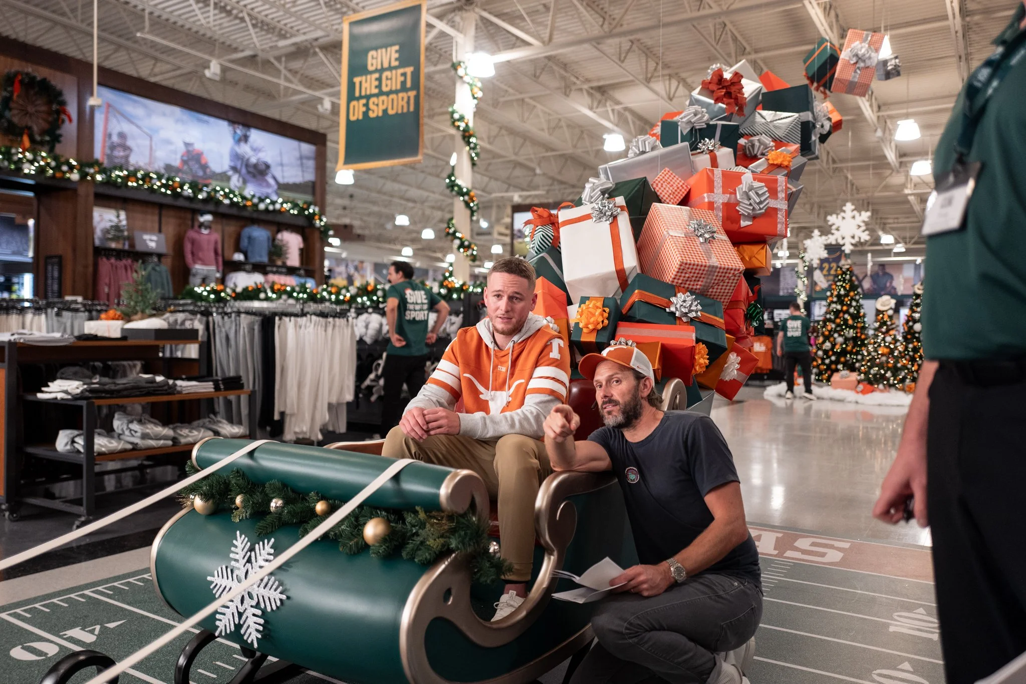 Two men sitting on a green sled in a sporting goods store decorated for Christmas, with a large Christmas tree and gift boxes in the background.