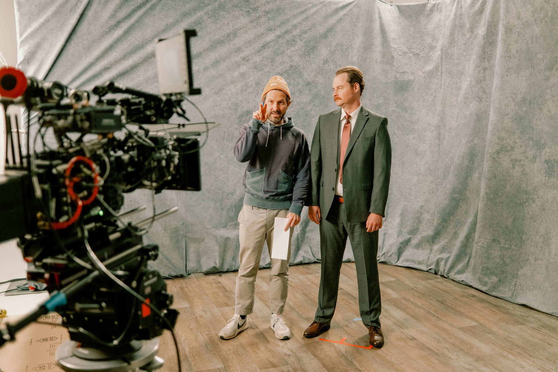 Two men stand in front of a gray backdrop on a film set, one in casual clothing and the other in a suit, with a camera set up facing them.
