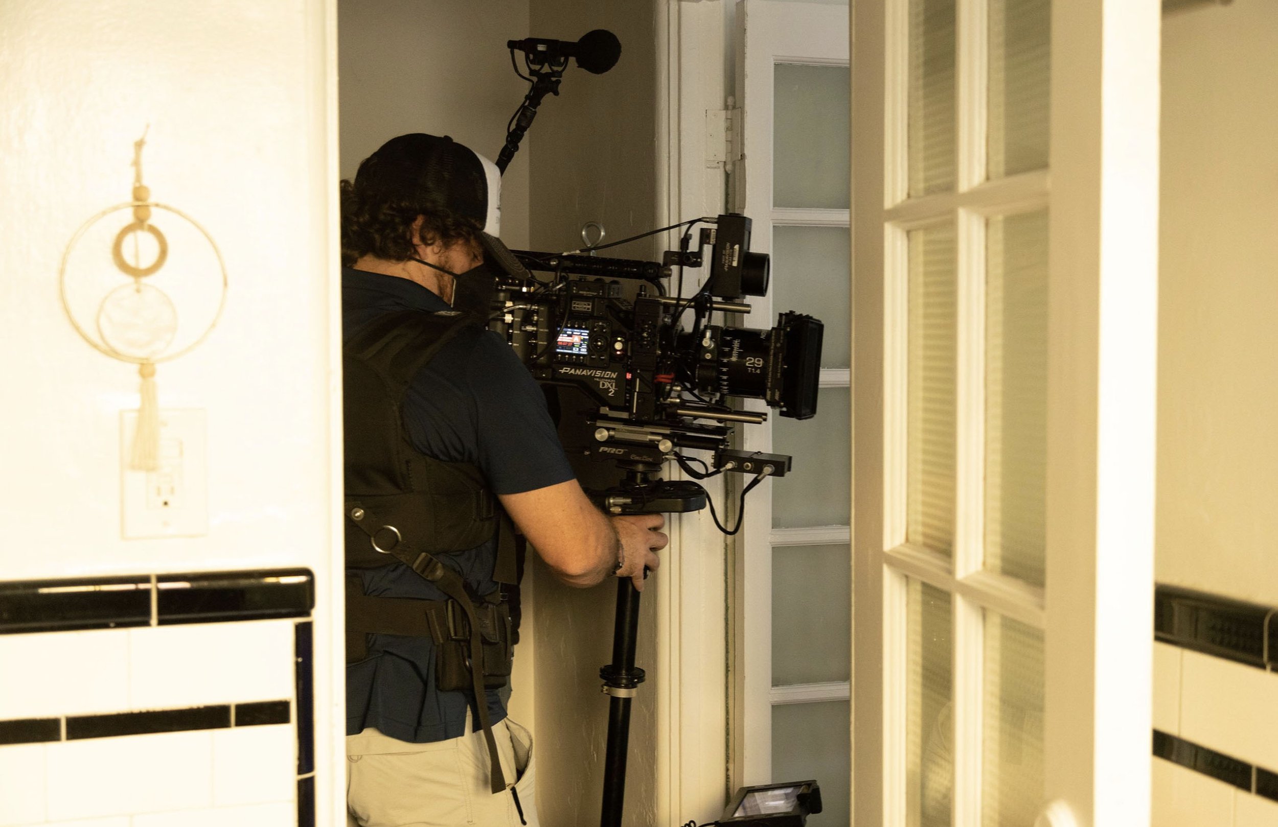A person wearing a baseball cap, black vest, and face mask operating a professional video camera on a set, with a doorway and glass-paneled door in the background.