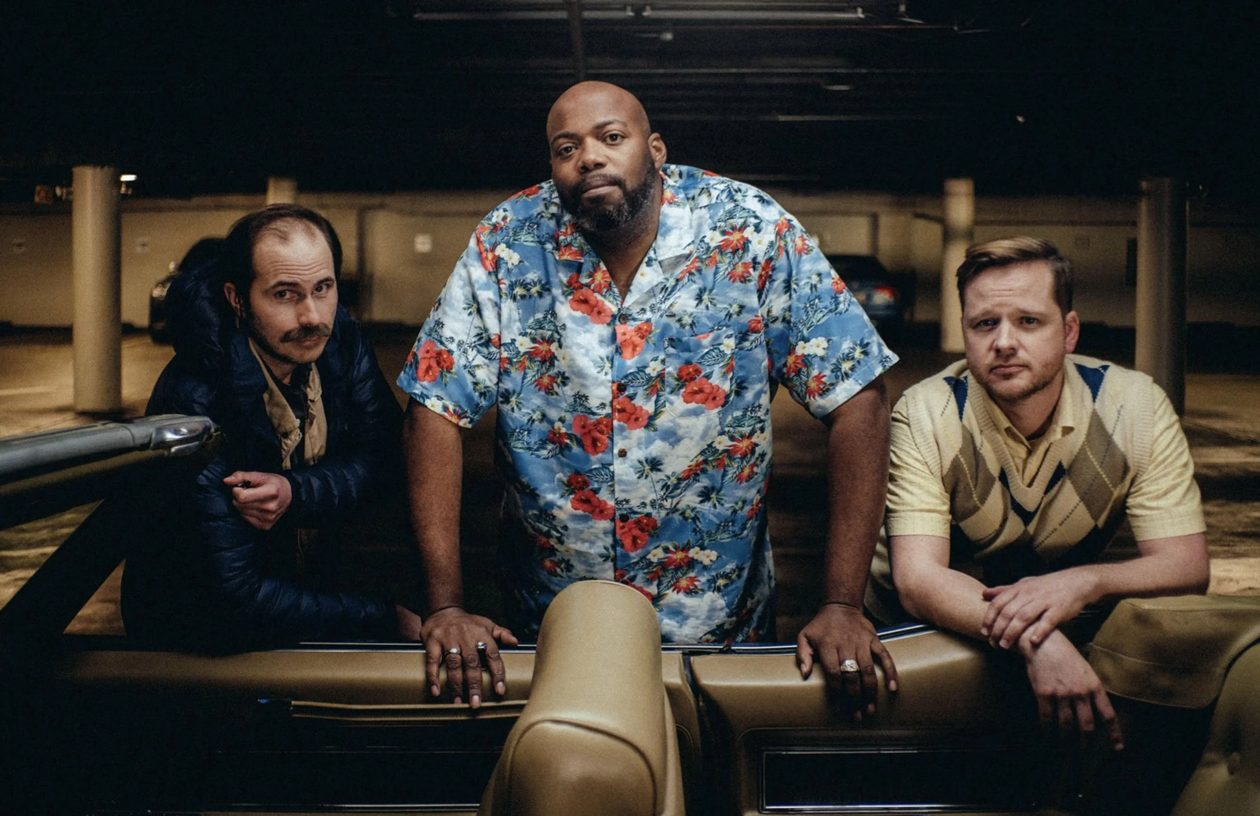 Three men in a parking garage, with the man in the center leaning on a car, wearing a blue floral shirt. The man on the left has long dark hair and a mustache, wearing a dark jacket. The man on the right has short hair, a beard, and is wearing a beig
