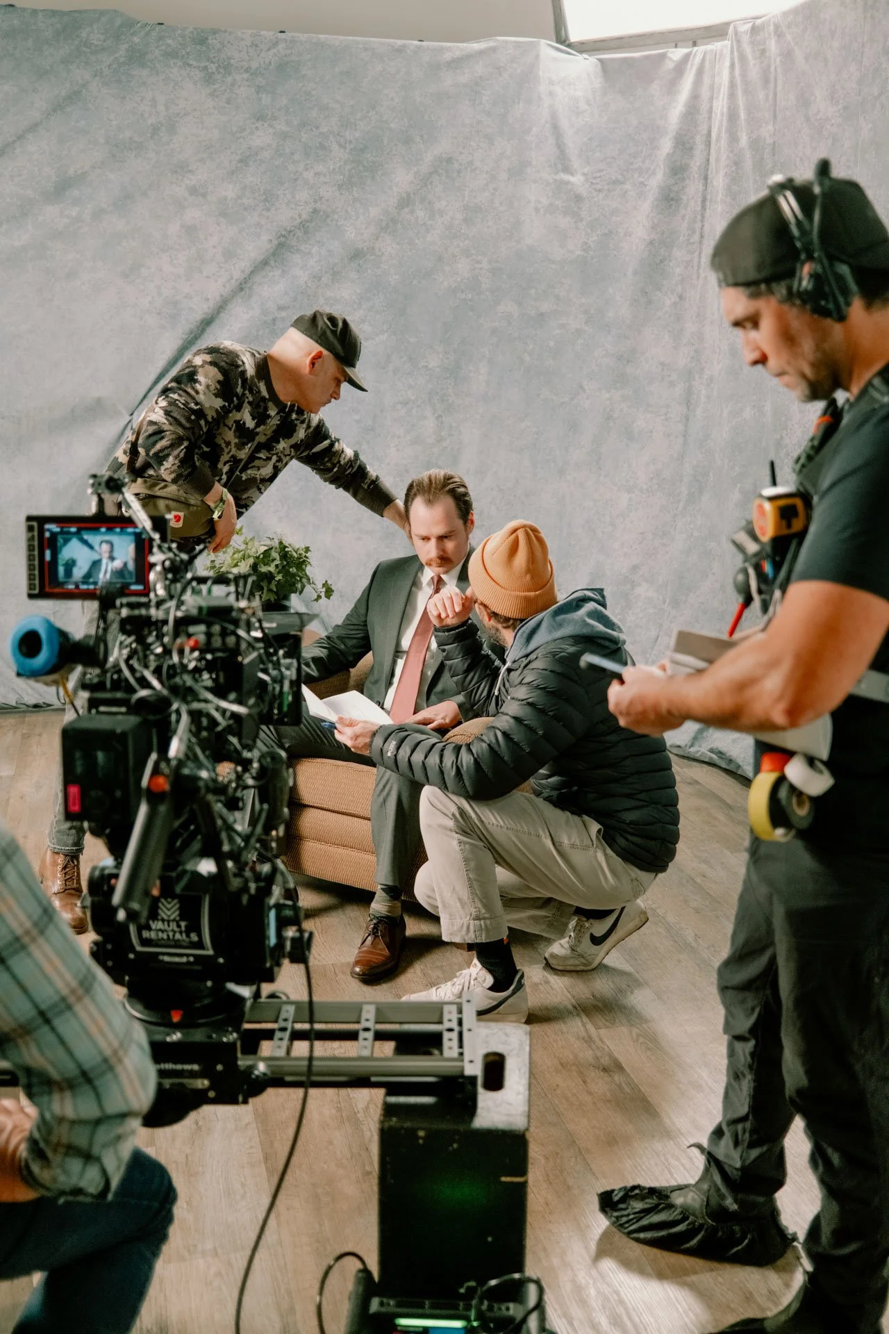 Film crew filming a scene with a man in a suit and another person in casual clothing, seated on a sofa, in front of a gray backdrop. Crew members are adjusting lighting and camera equipment.