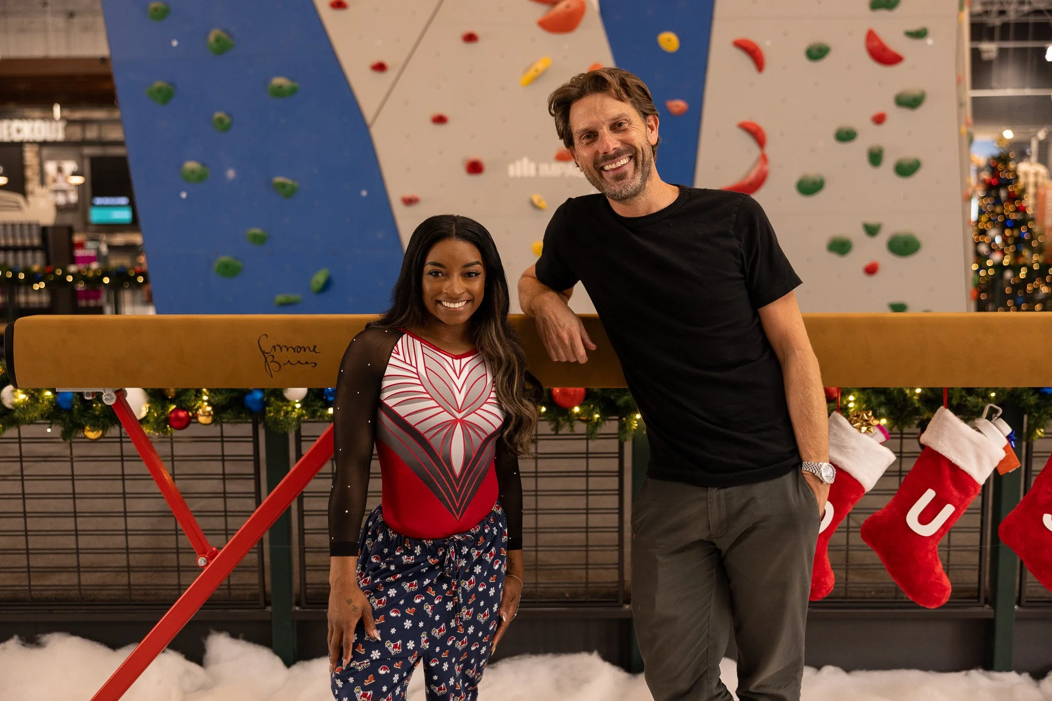 Two smiling people, a woman and a man, posing in front of a Christmas display with Christmas stockings and a decorated Christmas tree in the background. They are standing outdoors in front of a large indoor climbing wall.