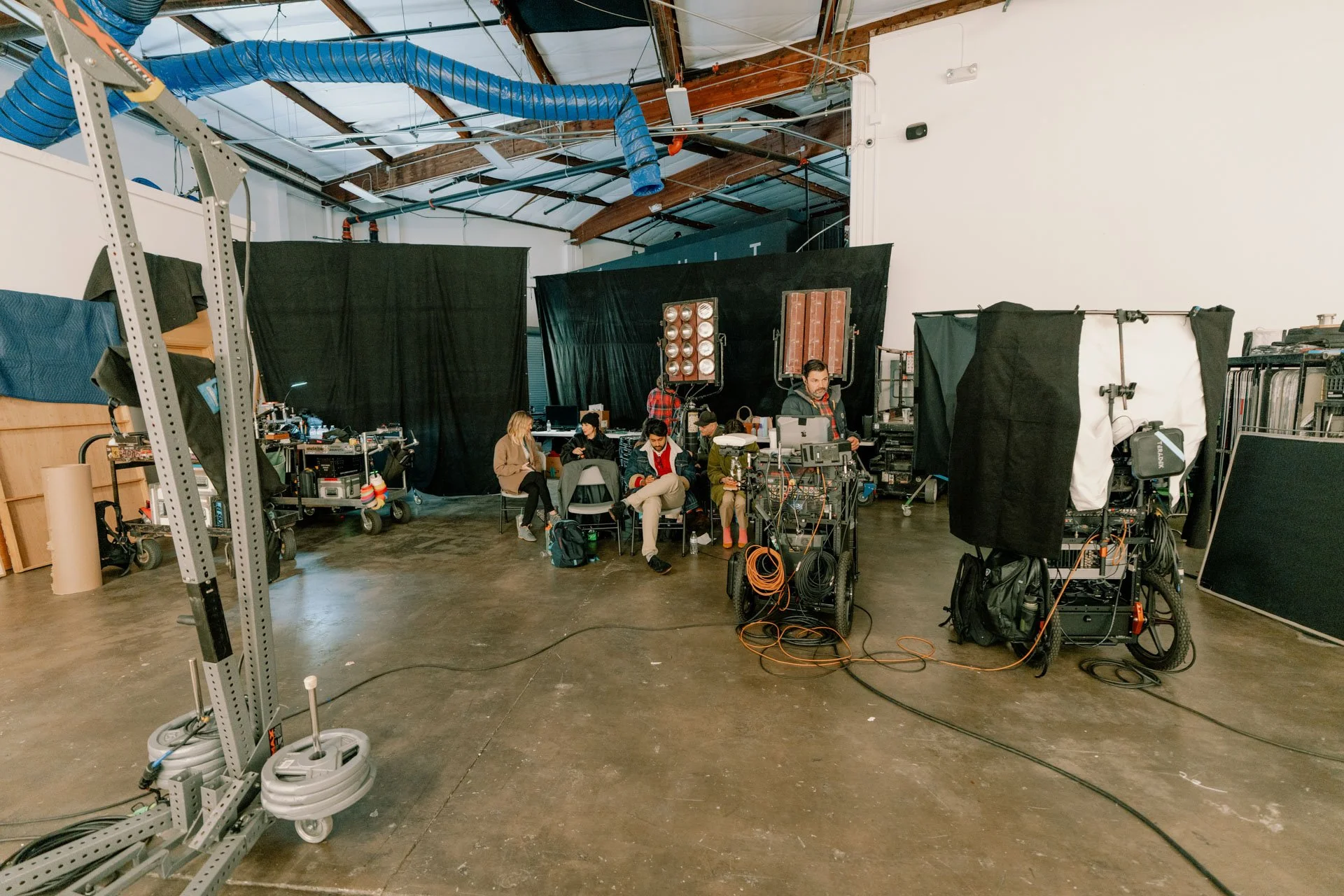 Behind-the-scenes view of a film or television production set with crew members working among equipment, black curtains, and lighting setups in a spacious industrial-style studio.