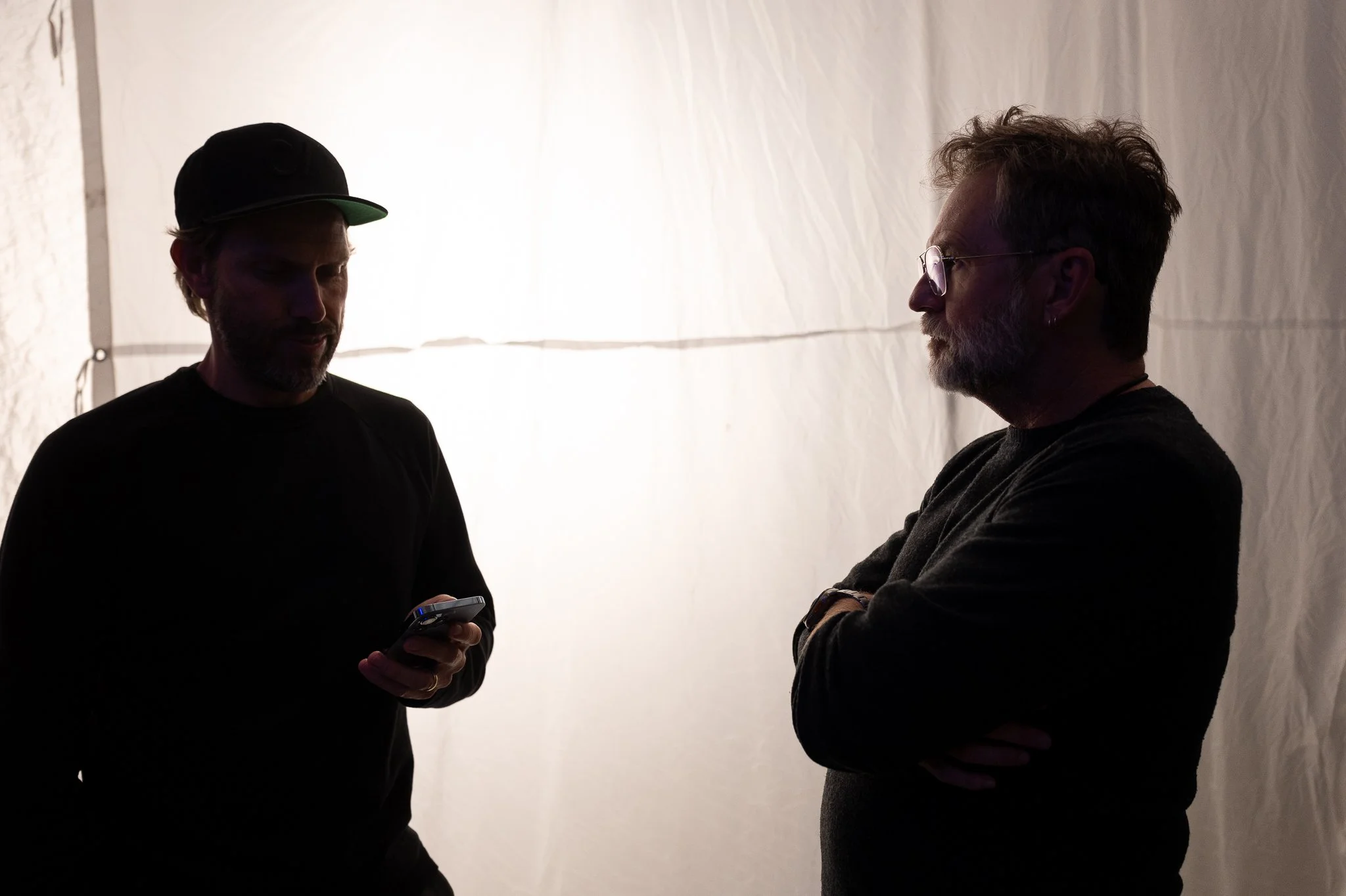 Two men standing in silhouette against a light background, one with a cap and holding a phone, the other with glasses and crossed arms, engaged in conversation.