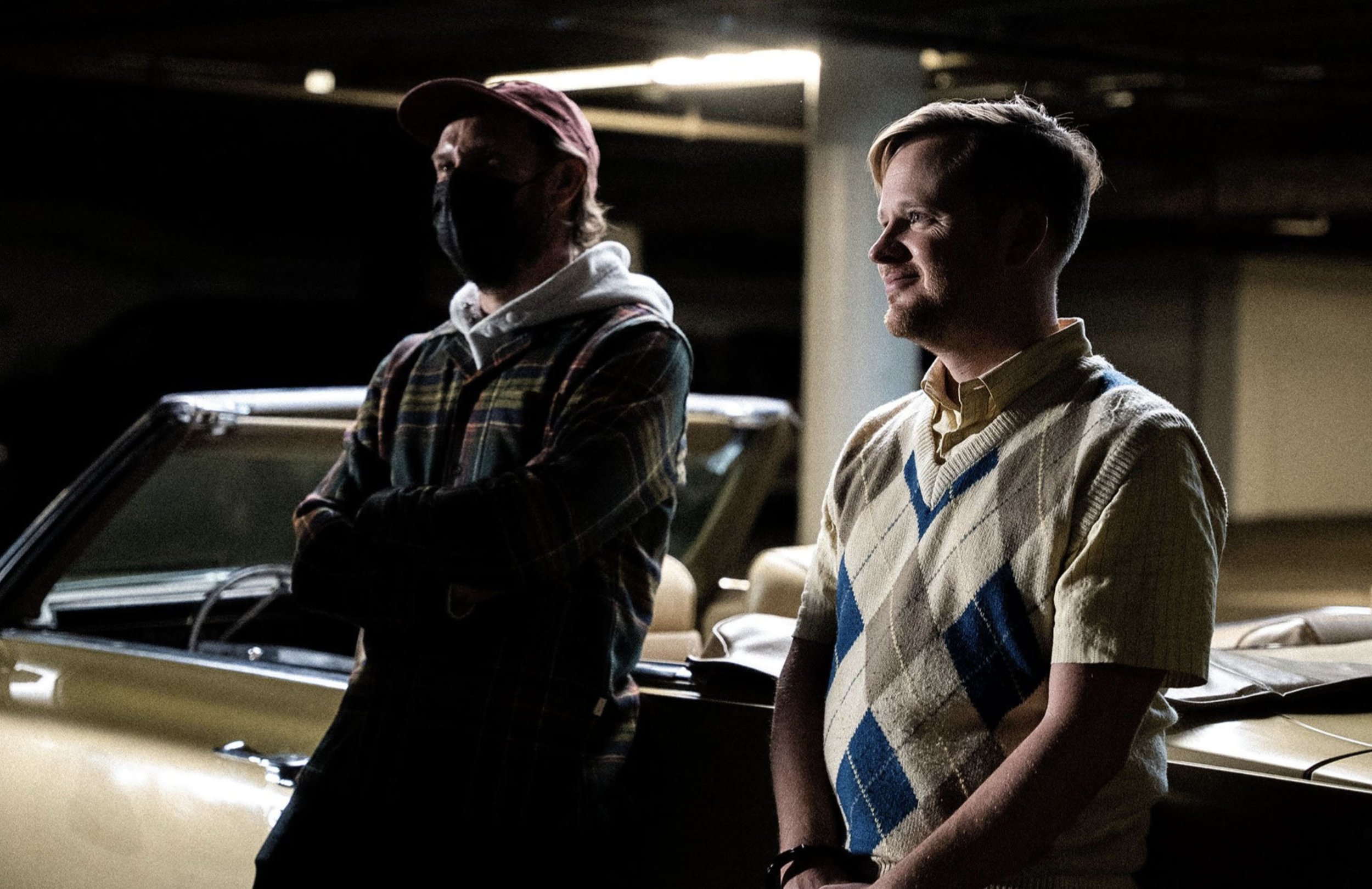 Two men standing in a dimly lit parking garage, one wearing a mask and a plaid jacket, the other smiling and wearing a short-sleeved sweater with an argyle pattern.