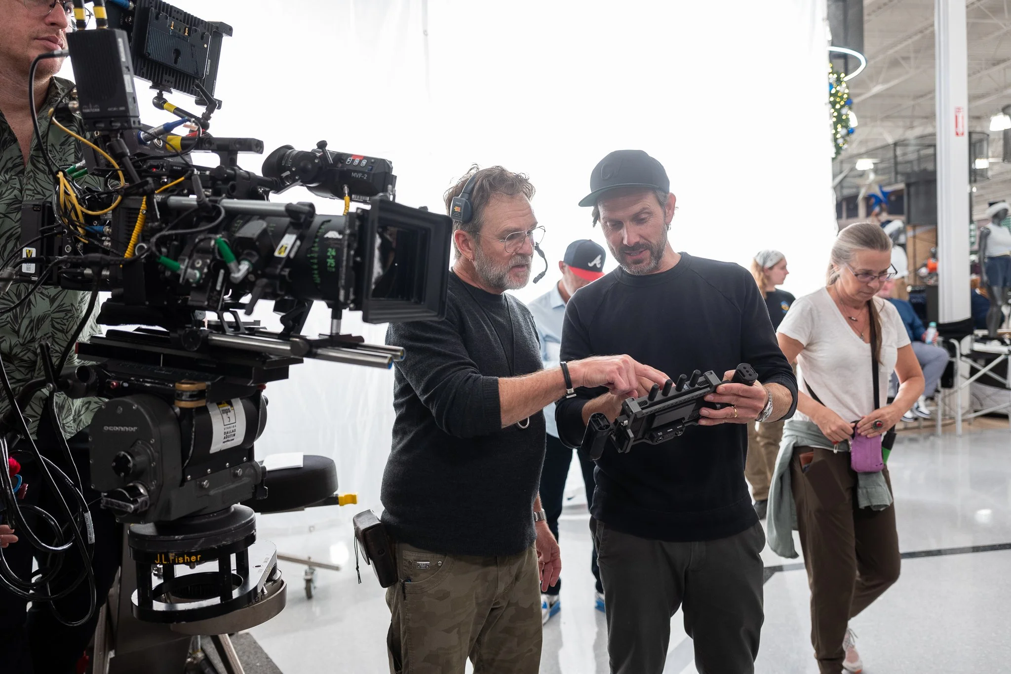 Film crew working on set with professional camera equipment, two men discussing and pointing at a device, other crew members in the background in a large indoor space.