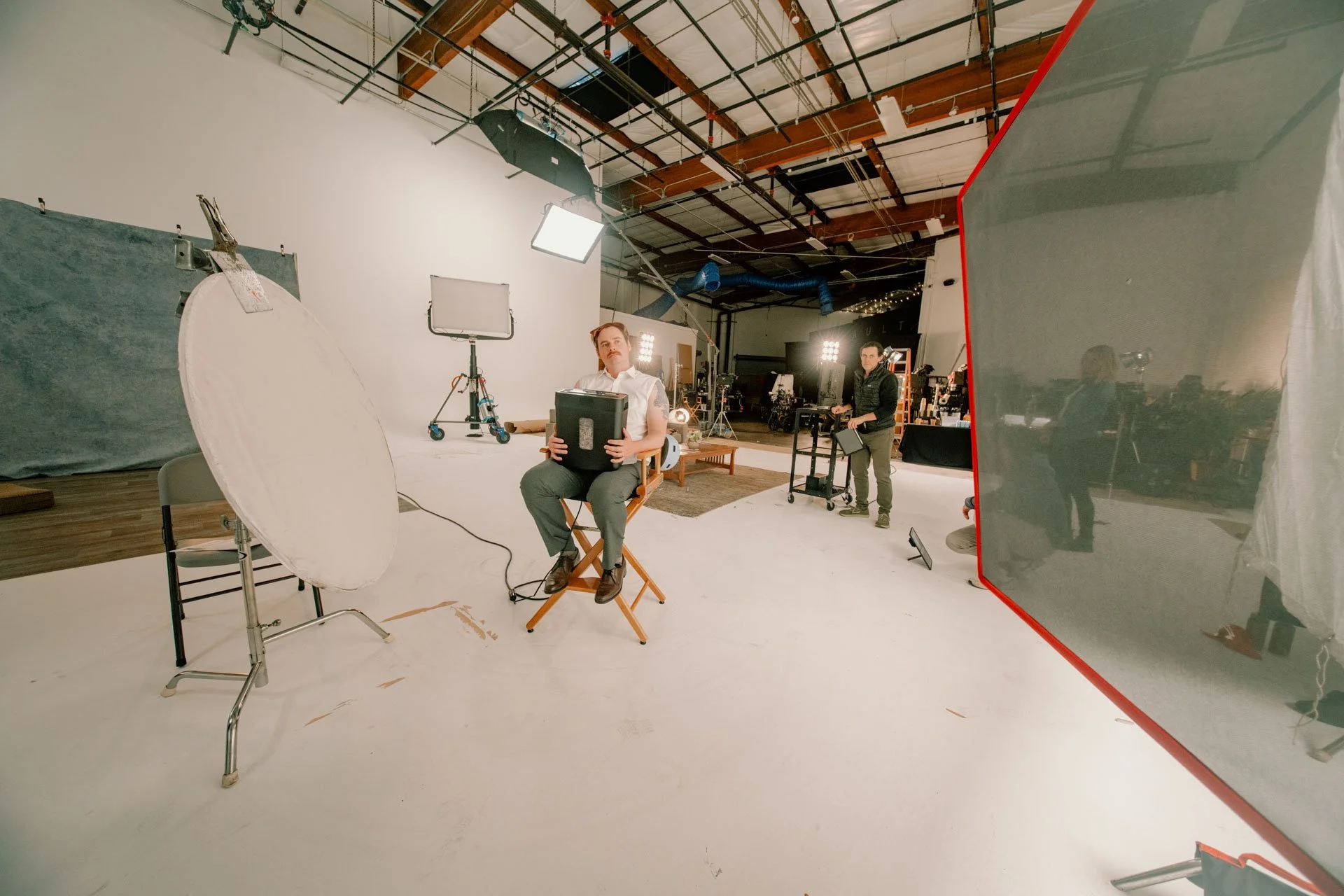 People in a photography studio, one sitting on a chair holding a black object and another operating a cart, surrounded by lighting equipment and a large diffuser, with a plain white background.