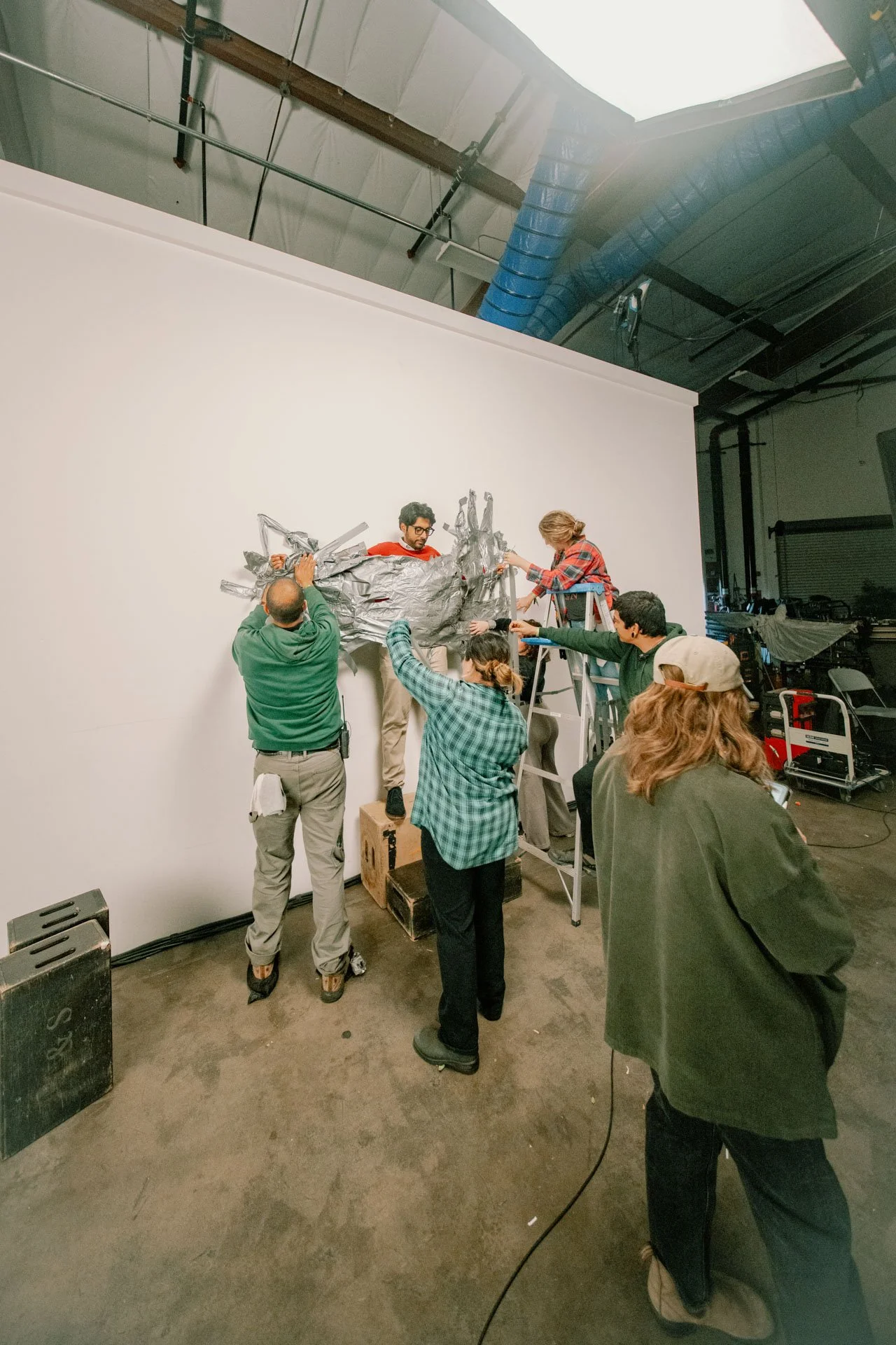 Group of people working together on an art or film project, with some applying metallic foil to a large sculpture or set piece on a white backdrop in an industrial studio setting.