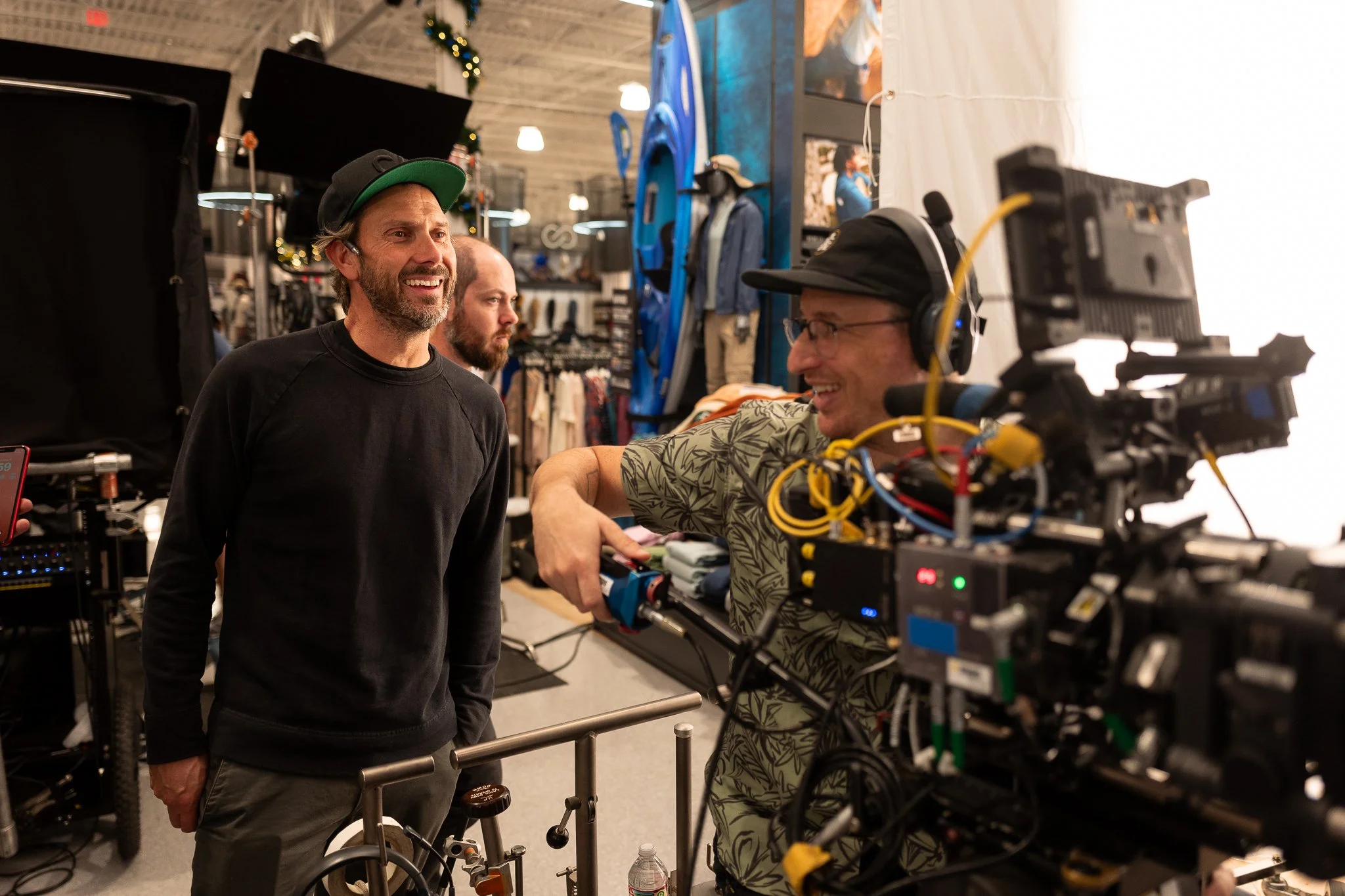 Two men are on a film set, one standing and smiling, the other sitting and operating camera equipment. The setting is a retail store with clothing and accessories in the background.