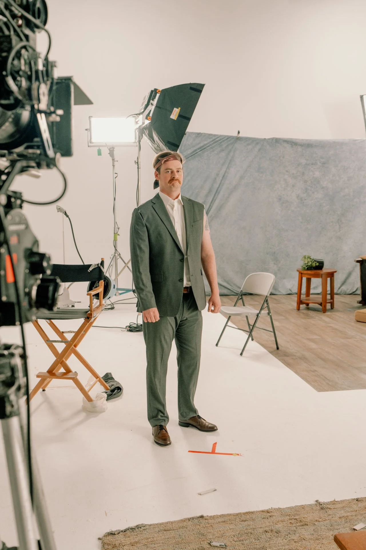 A man with a mustache, dressed in a gray suit with a torn sleeve, standing in a film studio with filming equipment, a chair, and a small table with a plant in the background.