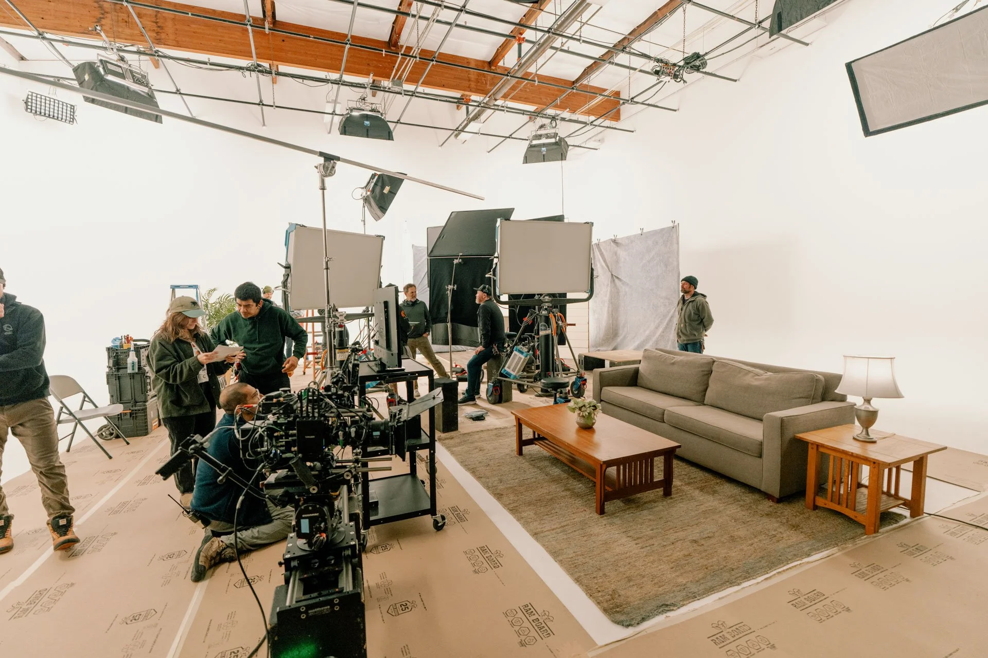 A film or television production set featuring a sofa and coffee table arranged as a living room scene. Crew members and equipment, including cameras and lighting, are set up around the set. Some crew members are inspecting or preparing equipment, wit