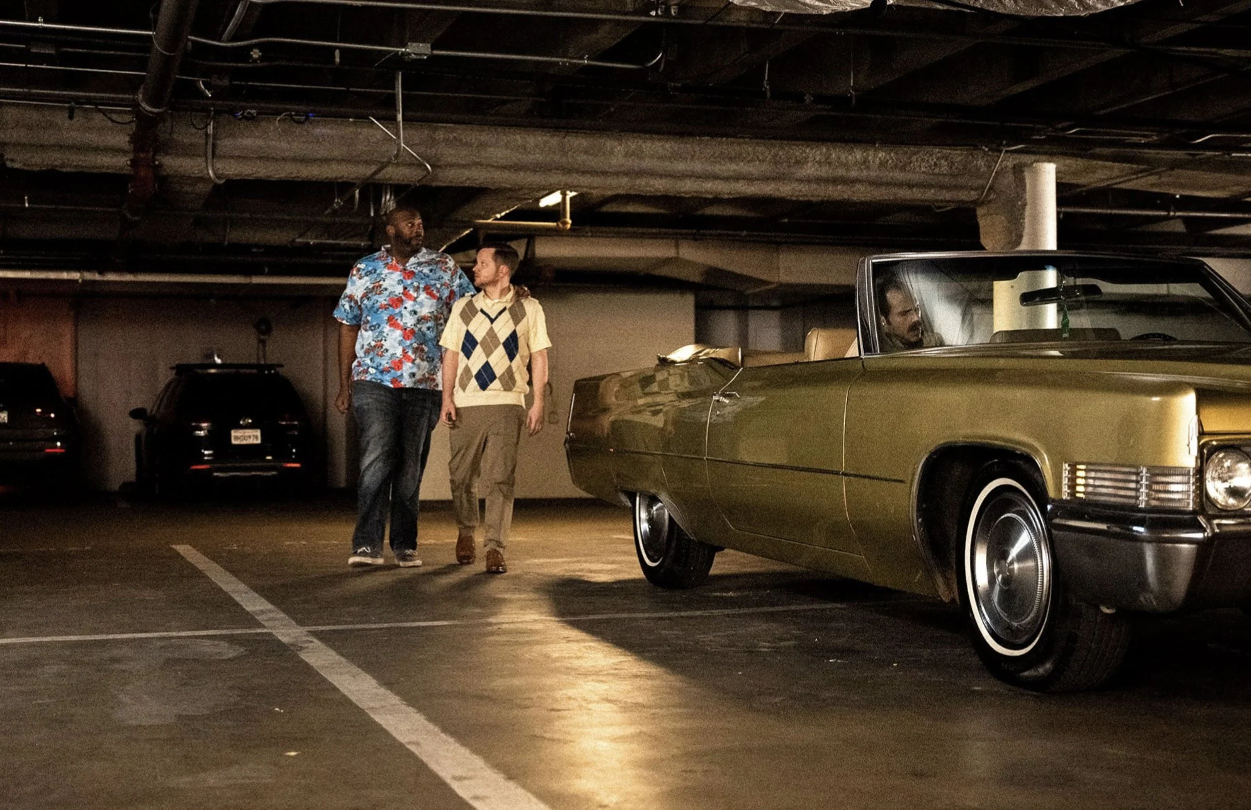 Two men are walking in a parking garage near a vintage yellow convertible car, with one man sitting inside the car.