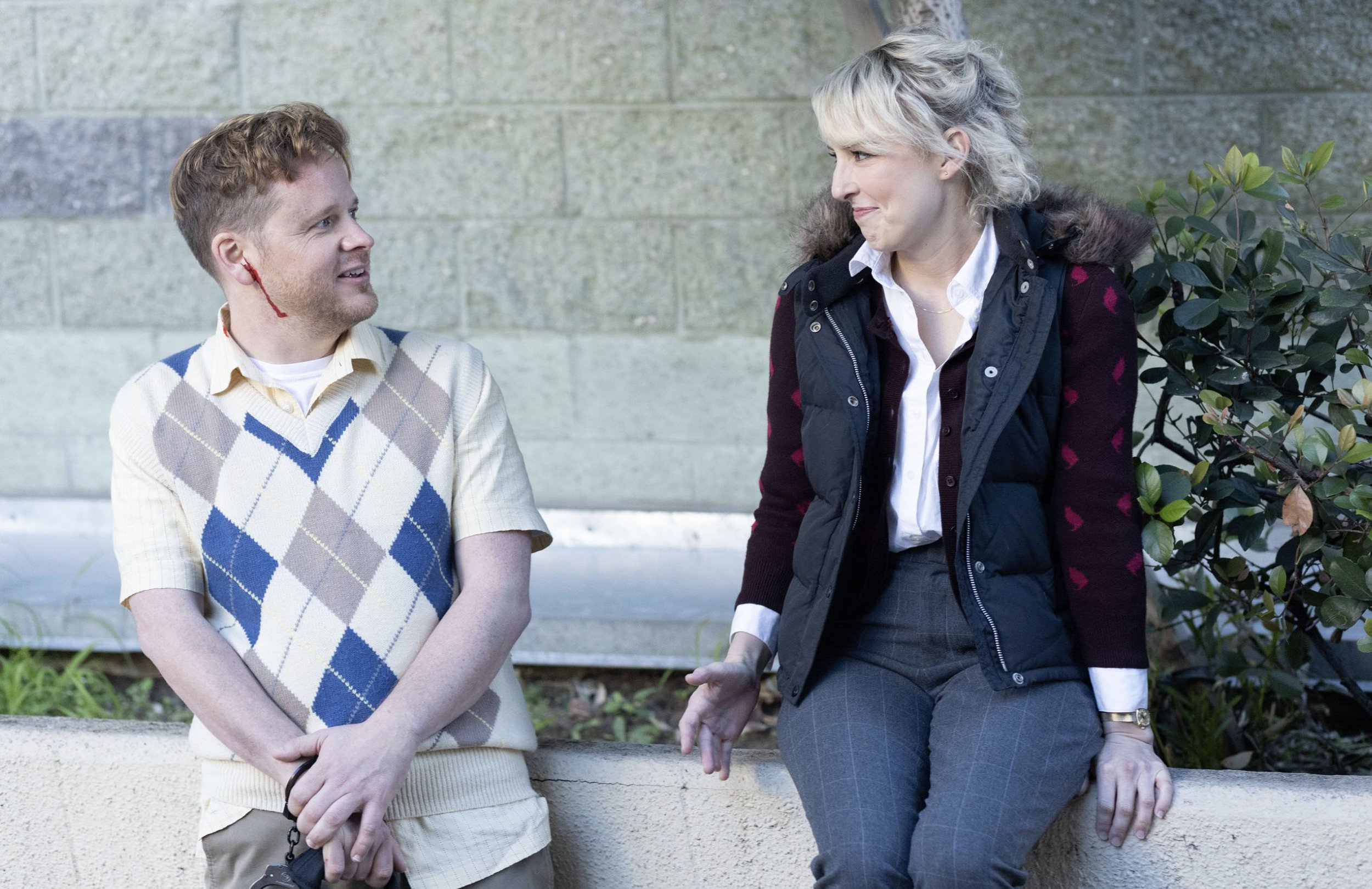A young man with a fake bloodstain on his face talking to an older woman sitting on a ledge outdoors near plants.