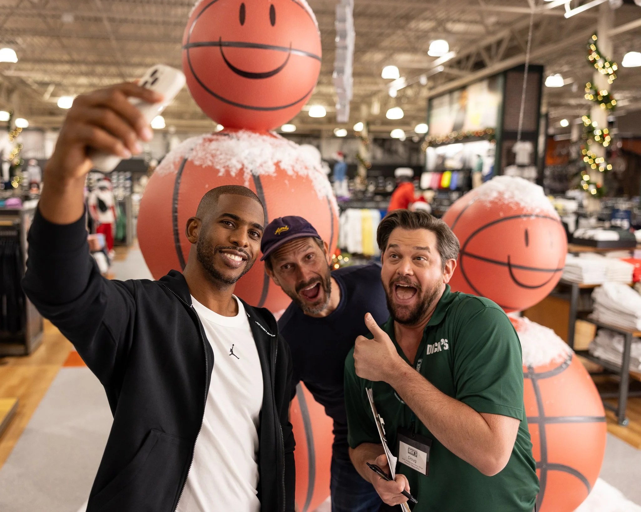 Three men taking a selfie in a retail store decorated with large basketball-shaped ornaments and Christmas decorations.