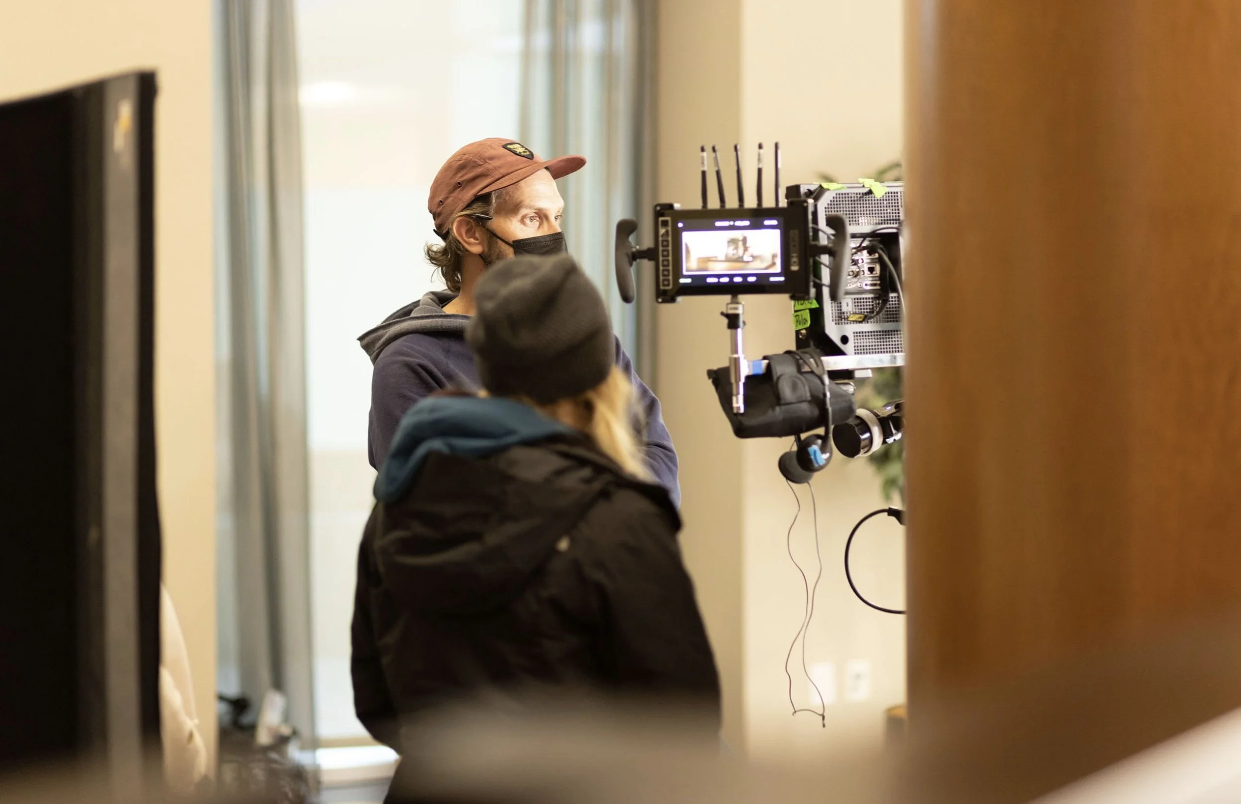 Two crew members, one wearing a face mask and a baseball cap, operating a camera on a film set indoors.