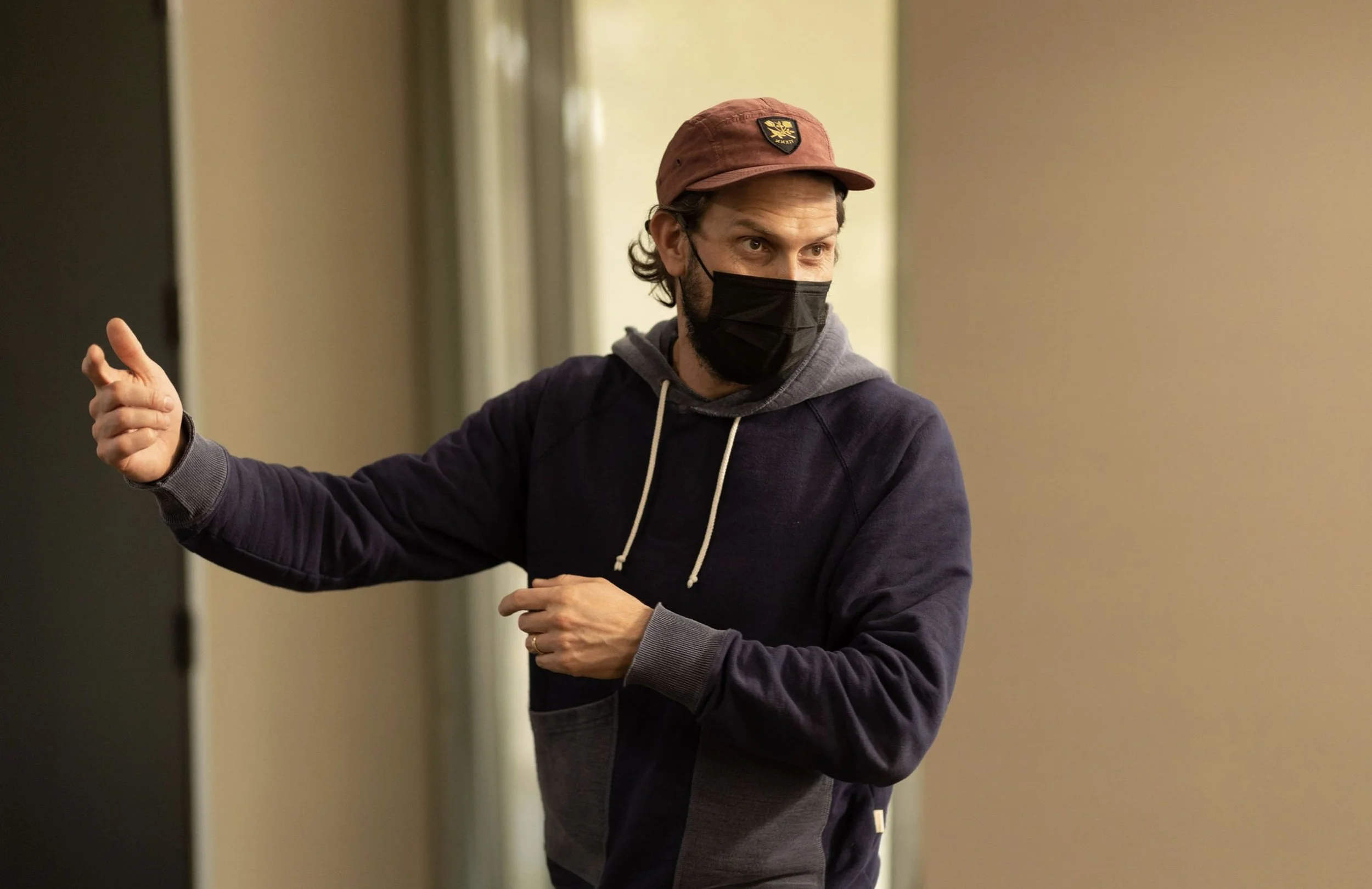 A man wearing a black face mask, a maroon baseball cap, and a dark hoodie, gesturing with his right hand and looking to his left indoors.