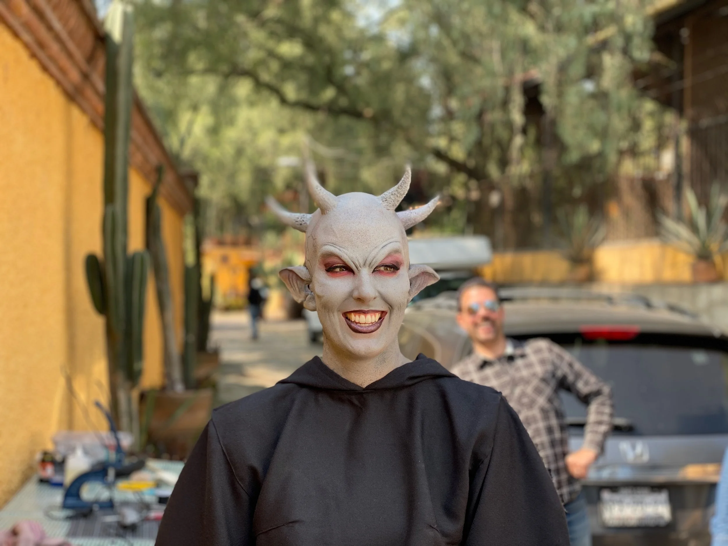 A person dressed as a mythical creature with white skin, horns, pointed ears, and exaggerated makeup, smiling outdoors near a yellow wall with cacti and trees in the background. A man wearing sunglasses and a plaid shirt is blurred in the background,