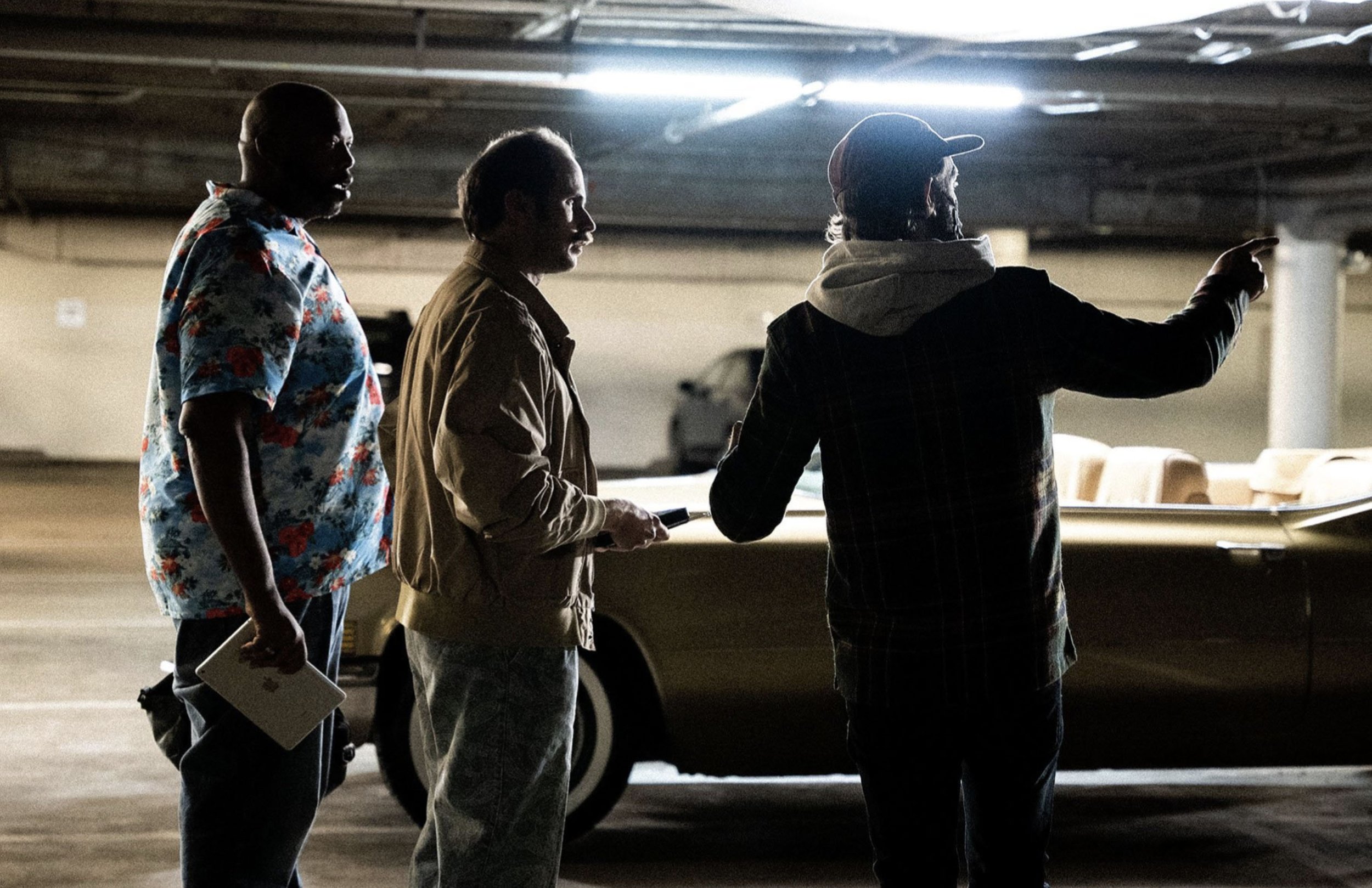 Three people standing in a parking garage, engaged in conversation, with one person actively pointing, the others listening, and a vintage convertible car nearby.