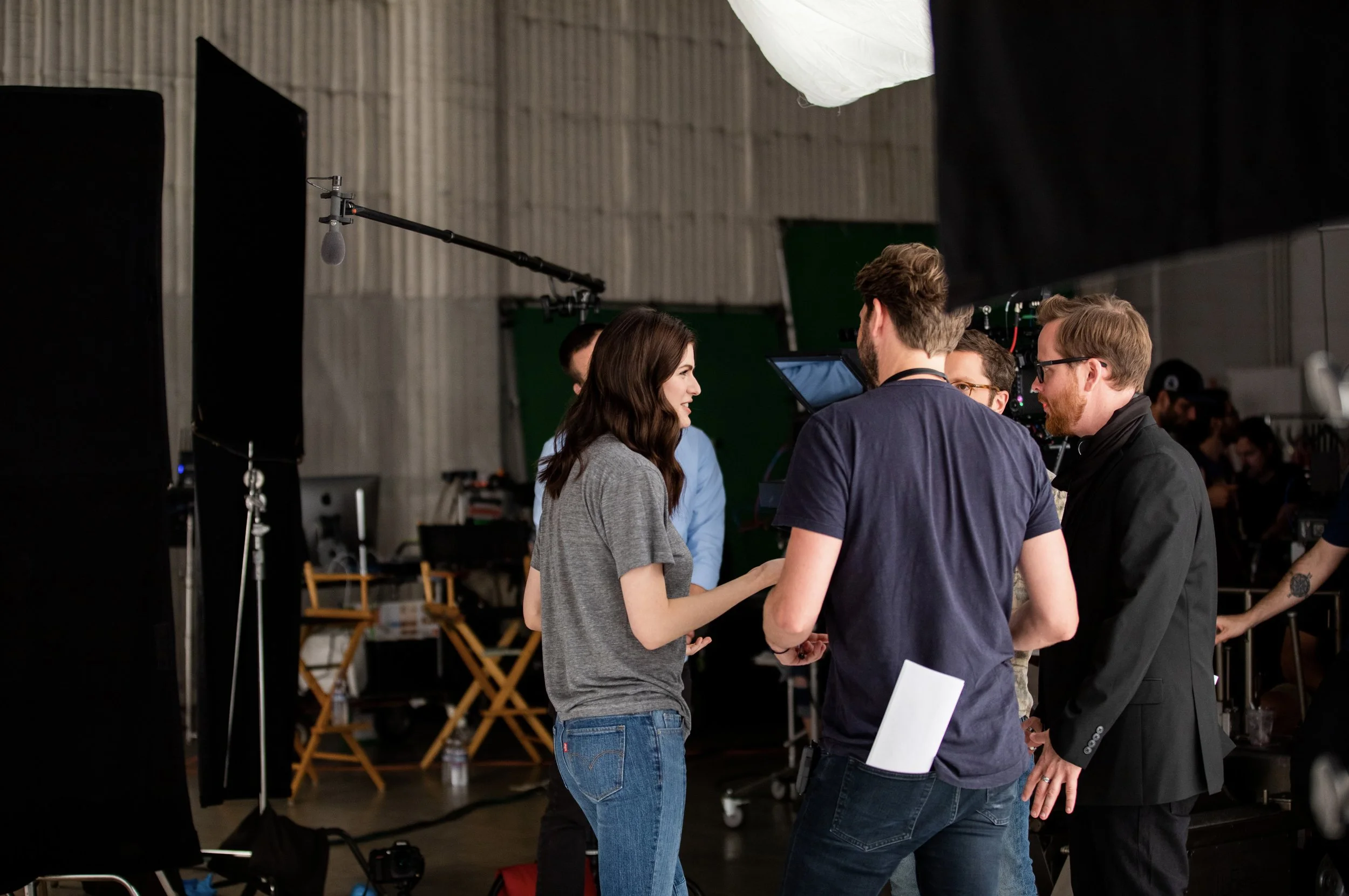 A group of people having a conversation on a film or television set with equipment around, including a boom mic, cameras, and lighting, in an indoor studio environment.