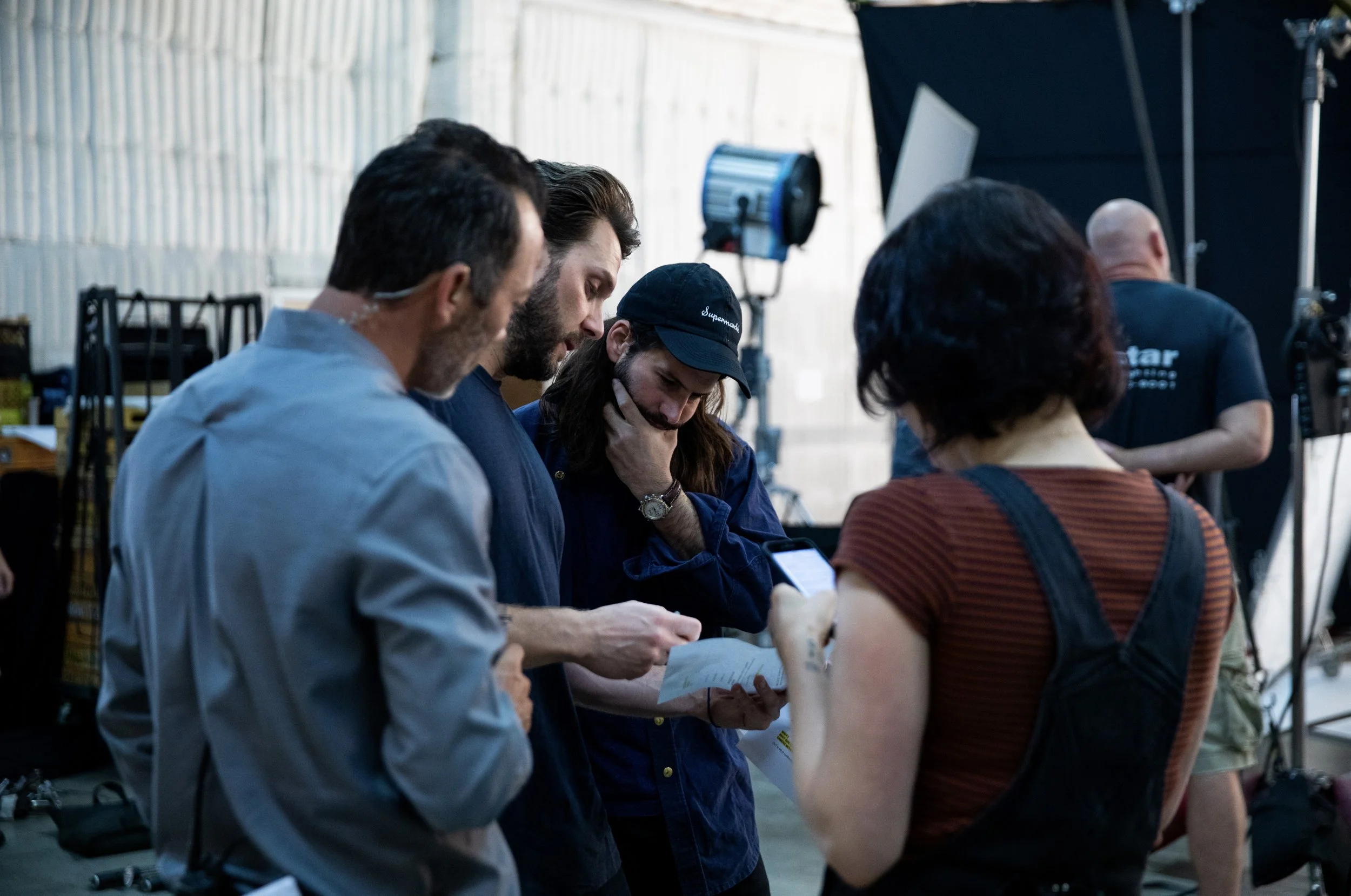 Group of people gathered indoors, standing closely together, some looking at their phones or a piece of paper, with photography equipment and lighting in the background.