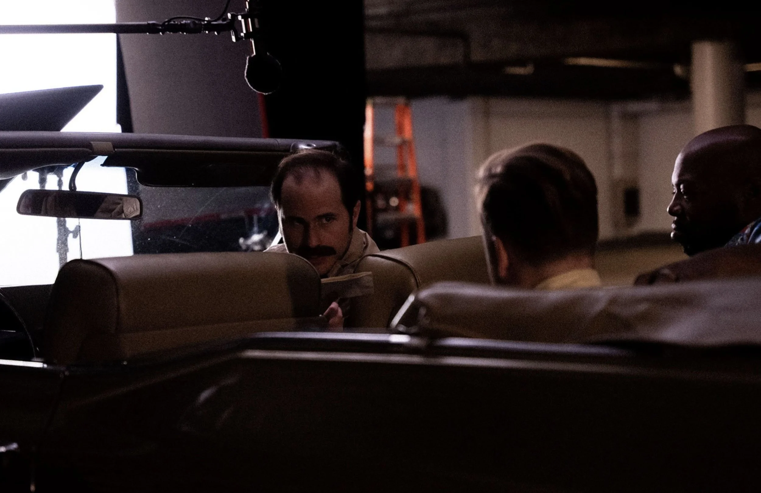 Three men sitting in a car on a film set, with one man looking directly at the camera, partially seen behind the car's interior, and a boom microphone overhead.
